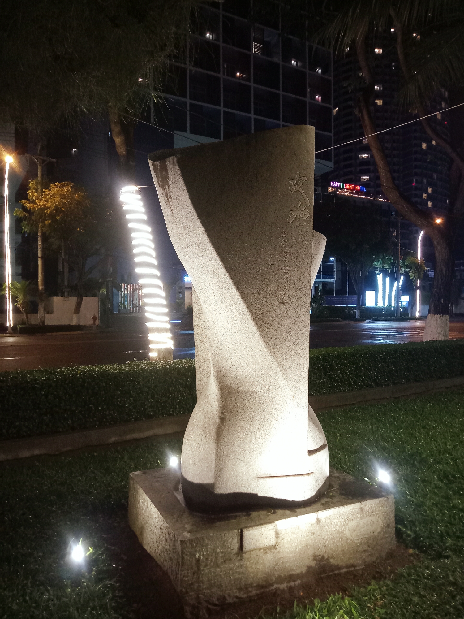 Captured at night in Nha Trang Ward, Vietnam, the scene is dominated by a stone sculpture placed on a raised stone platform on a grassy patch. Several small spotlights illuminate the base and the ground around the sculpture. A trimmed hedge runs parallel to the sculpture’s base. Beyond the hedge, a street is visible, featuring trees decorated with strings of lights that run vertically. Across the street, buildings are discernible, with one bearing the sign "HAPPY LIGHT HOTEL" illuminated in bright, multicolored lights.