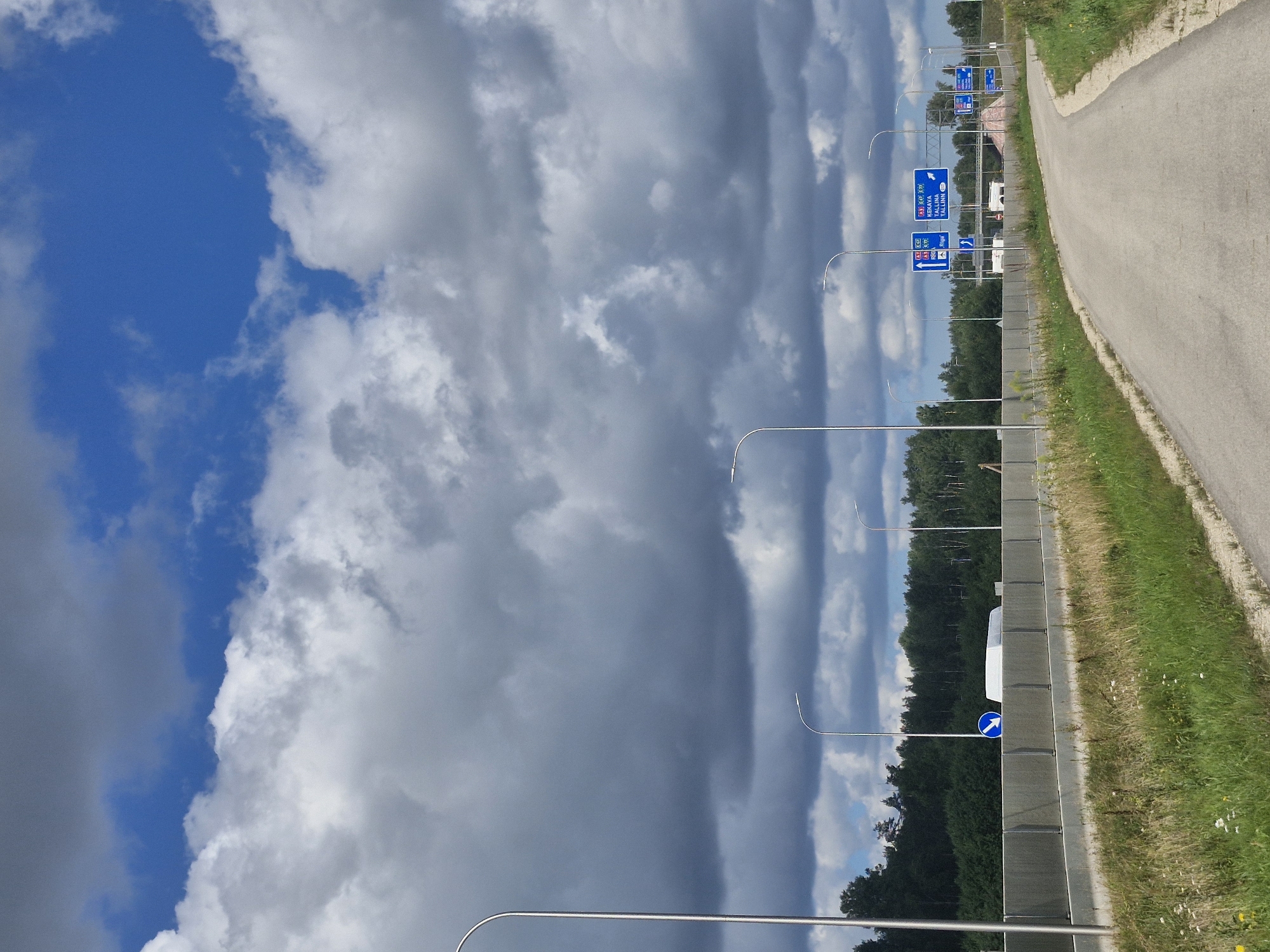 The image depicts a roadside scene with a paved path curving to the right. In the background, there is a highway with a concrete barrier and trees lining the distance. Several streetlights are visible along the highway. There are also road signs indicating directions to various locations. The sky is filled with dramatic clouds, with patches of blue visible. The environment appears to be rural or semi-rural, with dry grass and weeds growing along the side of the path. There are no people or animals visible in the frame. The scene is static and the lighting suggests it is daytime. This location is likely near Kursīši, Latvia, given the road signs indicating destinations within Latvia.