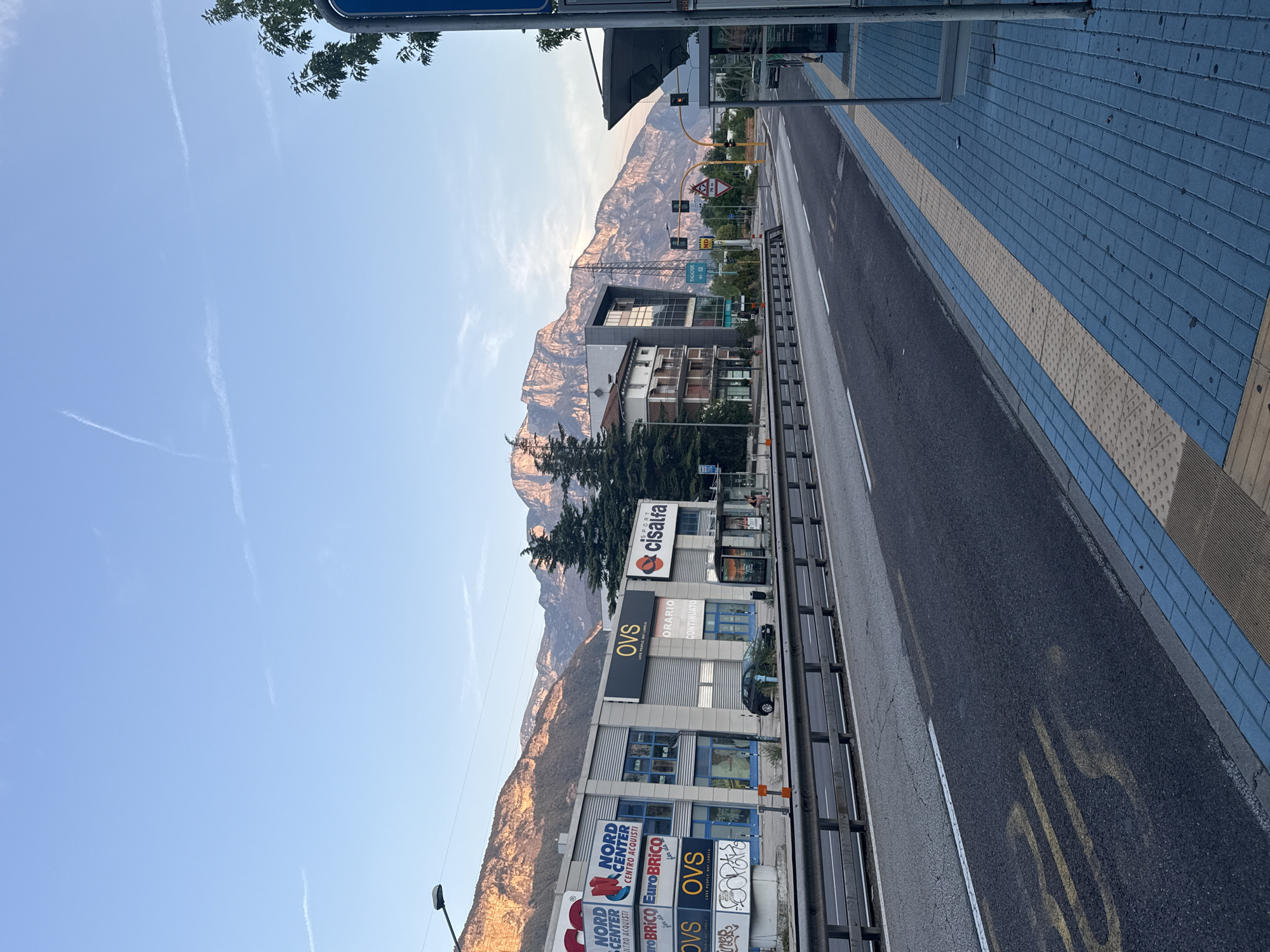 The image captures a street scene in Trento, Italy, likely during early morning or late afternoon given the lighting. A wide, paved road with lane markings runs diagonally through the frame, leading towards a mountainous backdrop. To the left of the road, a modern commercial building complex is visible, featuring signage for "NORD CENTER CENTRO ACQUISTI," "EuroBrico," and "OVS." The building has large glass windows and a metallic facade. A dark-colored car is parked in front of the building.
Adjacent to the road on the right is a bus stop shelter with a blue roof and metal supports. The sidewalk area leading to the bus stop is paved with blue and yellow bricks, incorporating tactile paving for the visually impaired. In the distance, traffic lights are visible, along with various signs. The imposing mountains, bathed in warm light, dominate the background. The sky is clear blue with faint white contrails. Several trees, including tall conifers, are scattered throughout the scene. A person is partially visible sitting at the bus stop. The overall impression is one of an urban periphery where commercial activity meets the natural landscape.