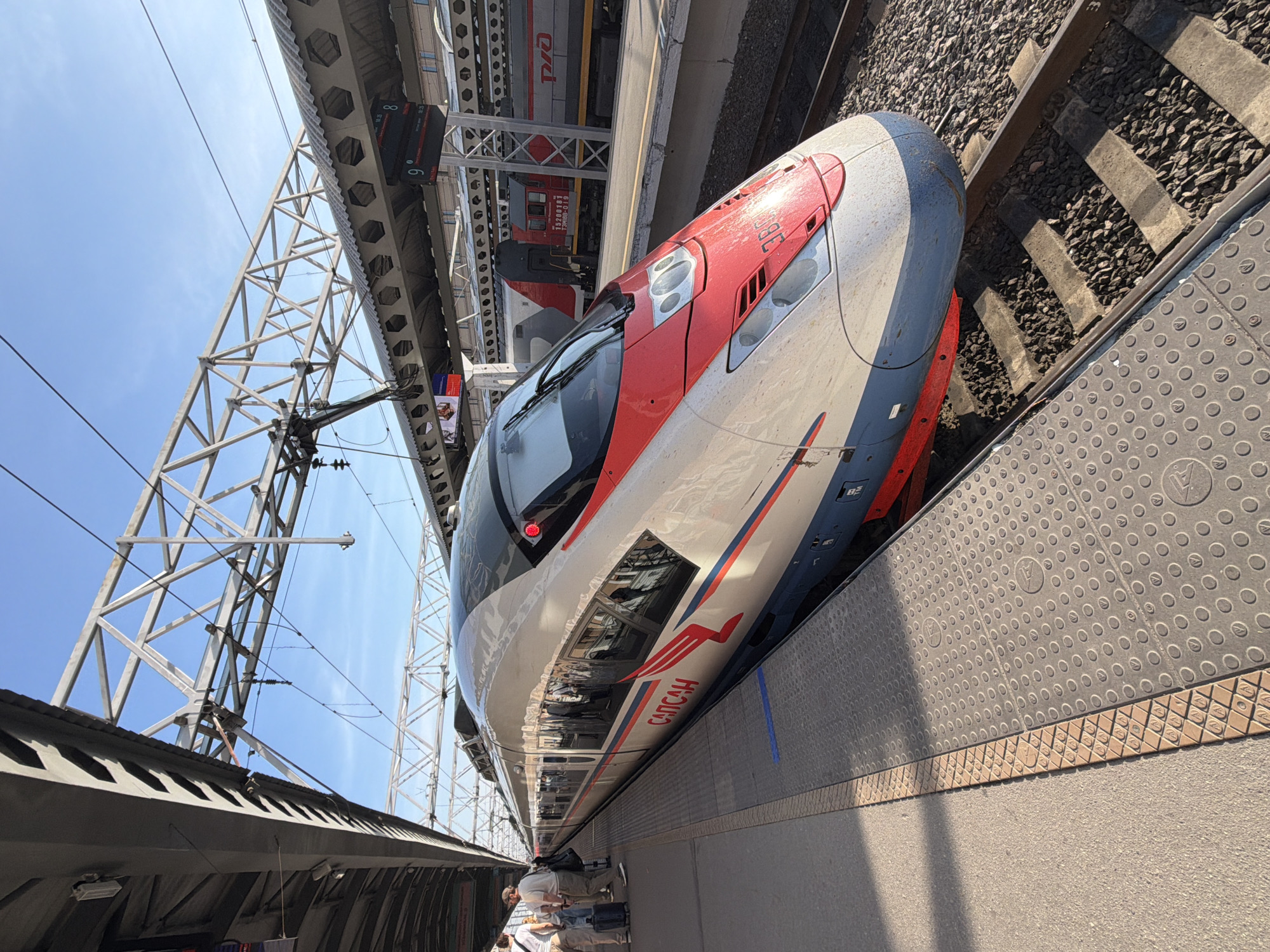 A high-speed train, identified by the Cyrillic text "САПСАН" and the number "307" on its front, is parked at a train station platform. The train is predominantly white with red and blue accents. Several people are visible on the platform, some standing near luggage, others near the train's entrance. The platform itself has a textured surface with a yellow tactile warning strip. Above the platform, a metal structure supports overhead electrical lines for the train. In the background, another train, red and grey with "РЖД" branding, is visible at a different platform, and train departure boards showing station numbers and times are mounted on the station's overhead structure. The sky is clear and blue, indicating daylight. The scene is likely at Moskovsky Rail Terminal in Saint Petersburg, Russia, a primary hub for Sapsan train services.