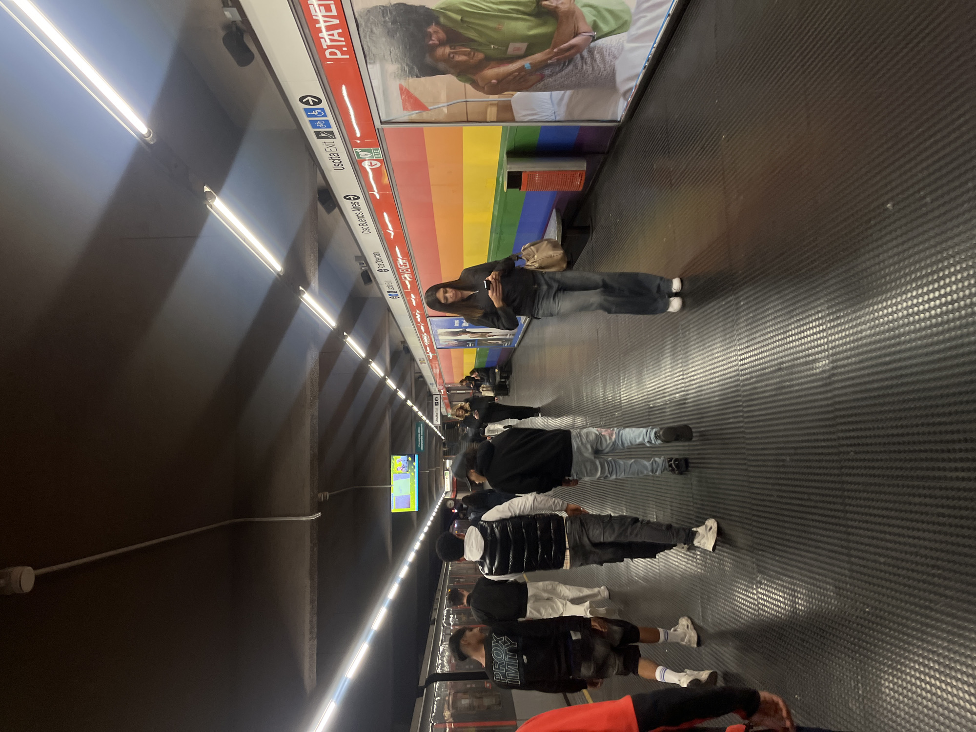 The image shows an underground train station platform in Milan, Italy. People are standing and walking along the platform, waiting for a train. The platform has a textured black floor and is lined with advertisements and station information. Overhead are fluorescent lights illuminating the scene. A sign with "P.TA VENEZIA" indicates the location is the Porta Venezia station. A woman stands near a poster with an embracing couple over a rainbow background, looking at her phone. Several other individuals are positioned in front of the train arriving at the station.