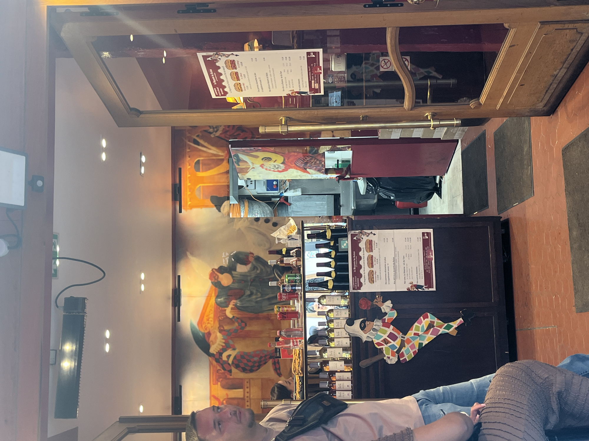 The image depicts the interior of a casual restaurant or cafe, likely located in Paris, France. The perspective is from a seated position, looking towards the entrance and a service counter. The photograph is rotated 90 degrees clockwise.
In the foreground, a man with short hair is visible, reclining on a bench or seat. He wears a light-colored top, jeans, and a black fanny pack across his chest. Part of another person's legs, also in jeans and a textured brown garment, can be seen to the right, suggesting they are seated next to him.
The background features a prominent service counter displaying various bottles, including wines (red and white), soft drinks like Coca-Cola and Sprite, and 1664 beer. A menu titled "La Baguette du Relais" is attached to the counter, listing items in French with prices, such as "Des pommes allumettes en plus" (fries extra) and "La Réserve vin du Tarn." A colorful, cutout figure of a harlequin is affixed to the counter. Behind the counter, a large, vibrant mural adorns the wall, depicting figures in what appears to be a Commedia dell'arte style.
The floor is tiled with reddish-brown, hexagonal tiles. Overhead, several spotlights illuminate the space. A speaker is mounted on a wall to the left. A glass door or entrance is visible, reflecting a "no smoking" sign and hinting at an exterior view. The presence of French text and the 1664 beer brand provide strong location cues for France, consistent with the Paris context. The overall atmosphere appears relaxed, with patrons enjoying the establishment's offerings.