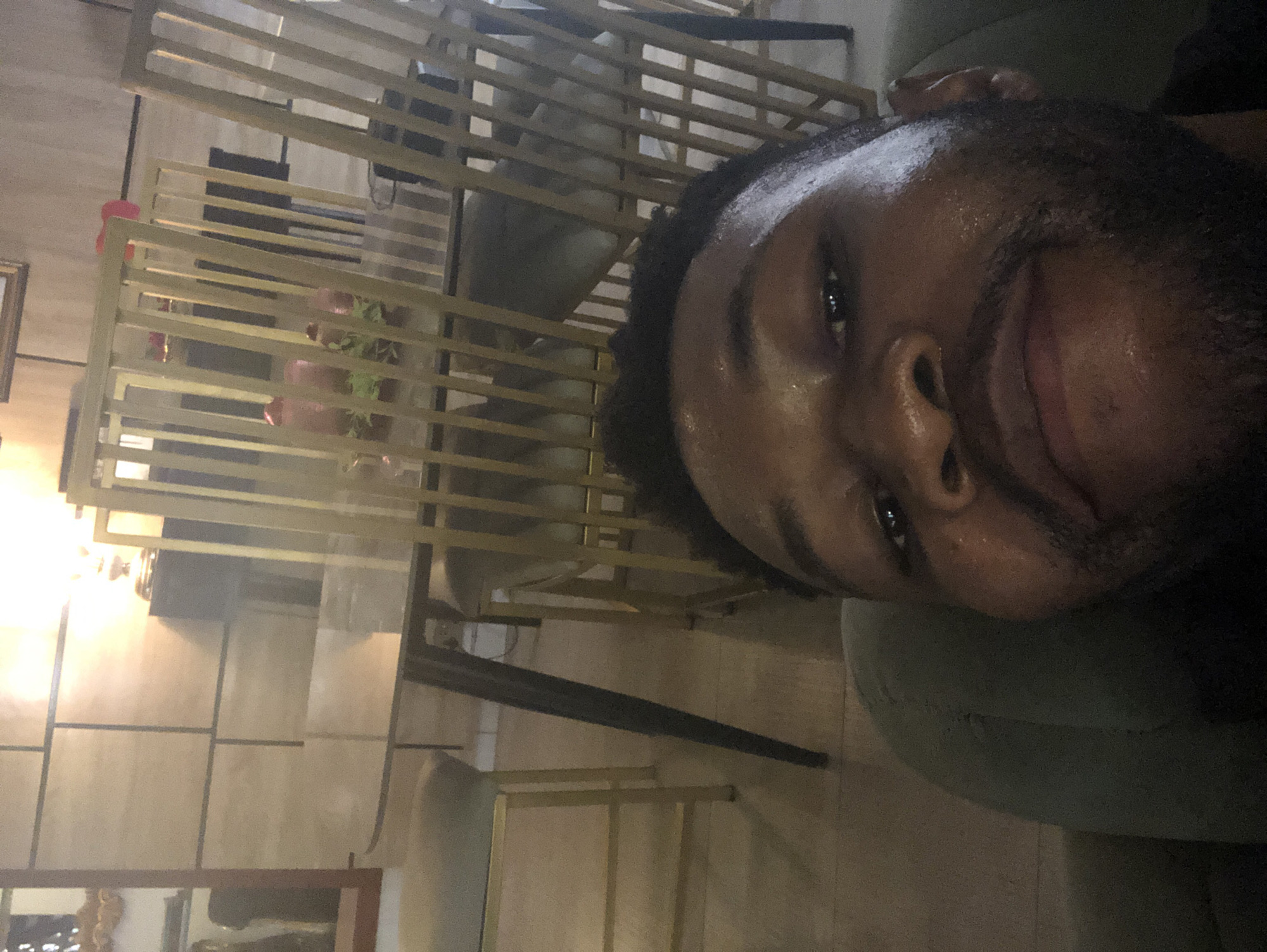 A close-up photograph shows the face of a smiling Black man, positioned against a blurred background that suggests an interior space. Visible behind him is a gold-colored structure with horizontal slats, possibly a decorative screen or partition. The background also shows chairs, plants in pots, and a wall with textured panels. The photograph does not provide enough information to determine a specific city or location.