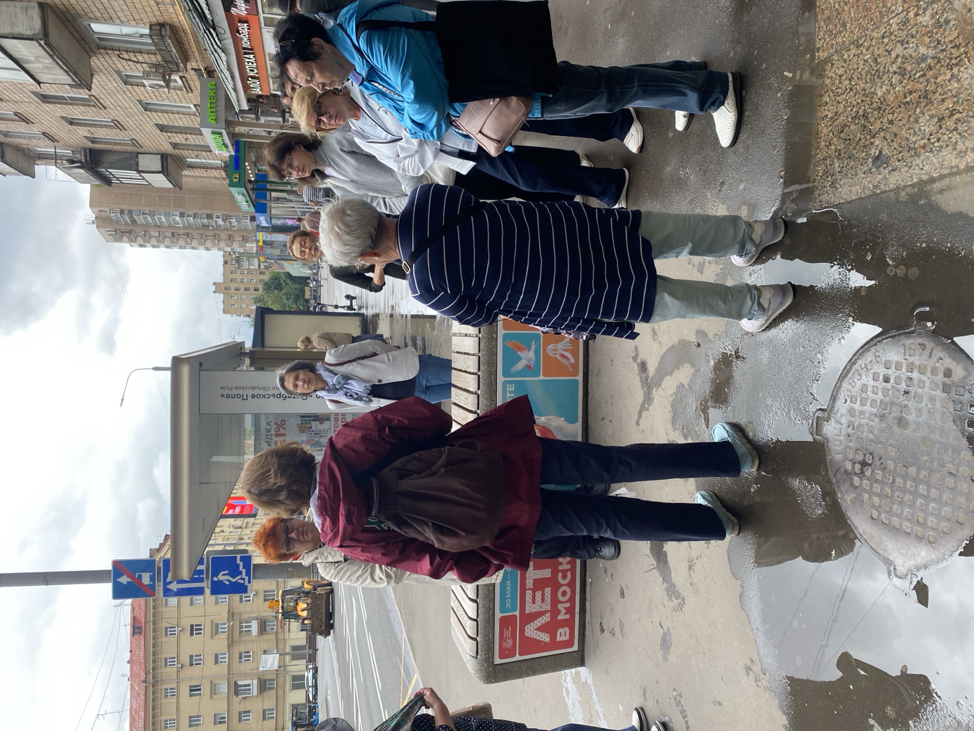 The image depicts a group of adults standing on a wet sidewalk in a city. In the background, there are buildings, a road with a yellow backhoe, and traffic signs. The foreground shows a bus stop shelter with a bench. The people appear to be engaged in conversation. The overall lighting suggests an overcast day.