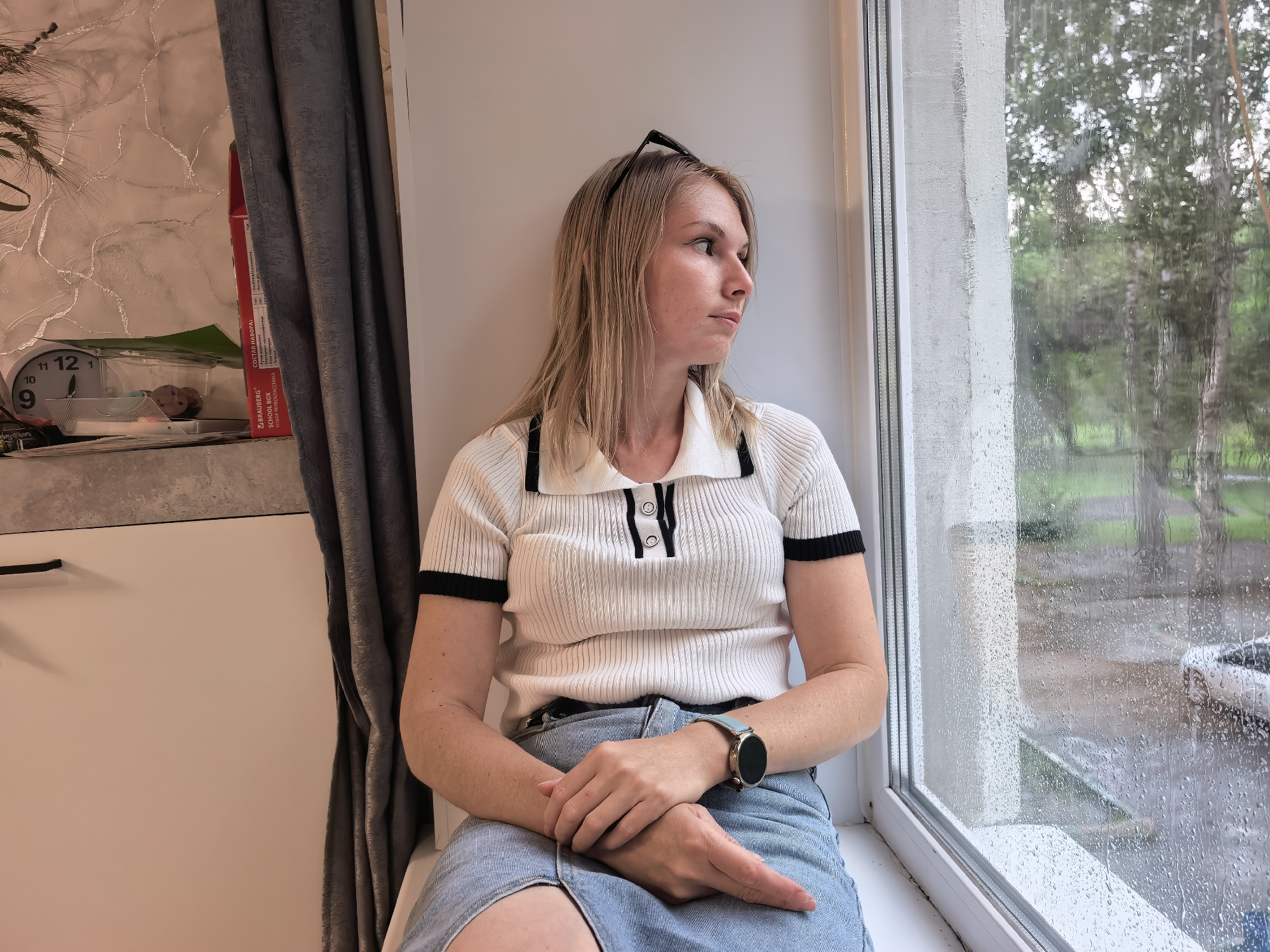 A young woman with blonde hair is sitting on a windowsill, looking out a rain-streaked window. She is wearing a white short-sleeved polo shirt with black trim and a light blue denim skirt. Her hands are clasped in her lap, and she is wearing a light blue smartwatch on her left wrist. Outside the window, trees and a parked car are visible through the raindrops. The interior of the room includes a gray curtain and a shelf with a clock and other items. The location is identified as Sterlitamak, Russia.