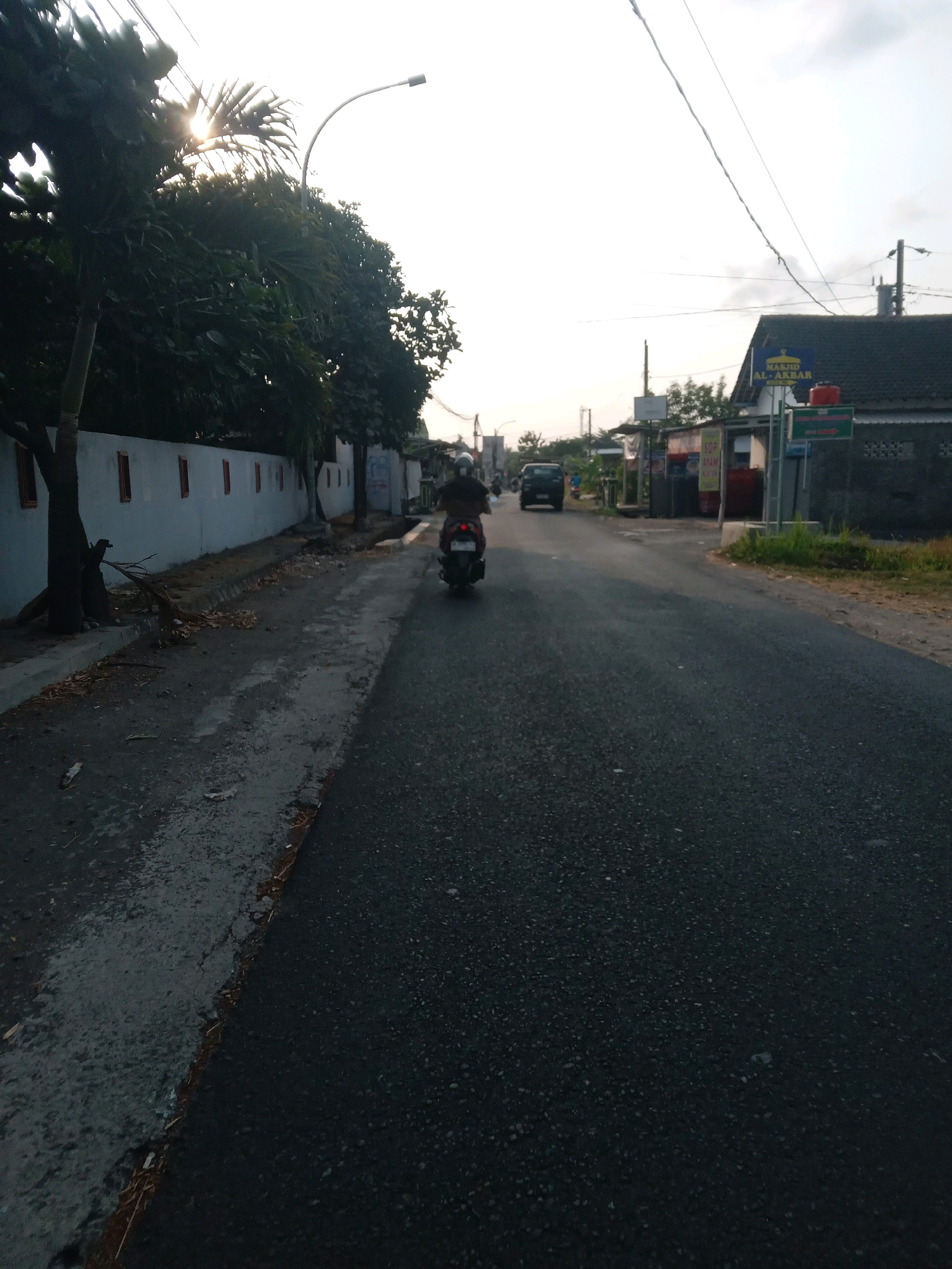 The image depicts a street scene in Banguntapan, Indonesia, likely during the early morning or late afternoon given the lighting conditions. A person is riding a dark-colored scooter down the asphalt road, facing away from the camera. The scooter has a red rear light visible. On the left side of the road, there is a low white wall with evenly spaced, rectangular openings or windows, bordered by trees and vegetation, some of which appear to be palm trees. A tall, curved streetlight is visible above the trees. On the right side of the road, there are buildings with dark roofs, including a building with a blue sign that reads "MASJID AL-AKBAR". There are also smaller structures that appear to be roadside stalls or shops, one with a sign indicating "SOP AYAM". Power lines stretch across the sky. The road surface is dark asphalt, with a lighter colored, possibly worn or unfinished, strip along the left shoulder. Debris, including leaves and small branches, is present on the shoulder. In the distance, another dark-colored vehicle is visible on the road. The sky is overcast with some areas of brightness, suggesting either sunrise or sunset.