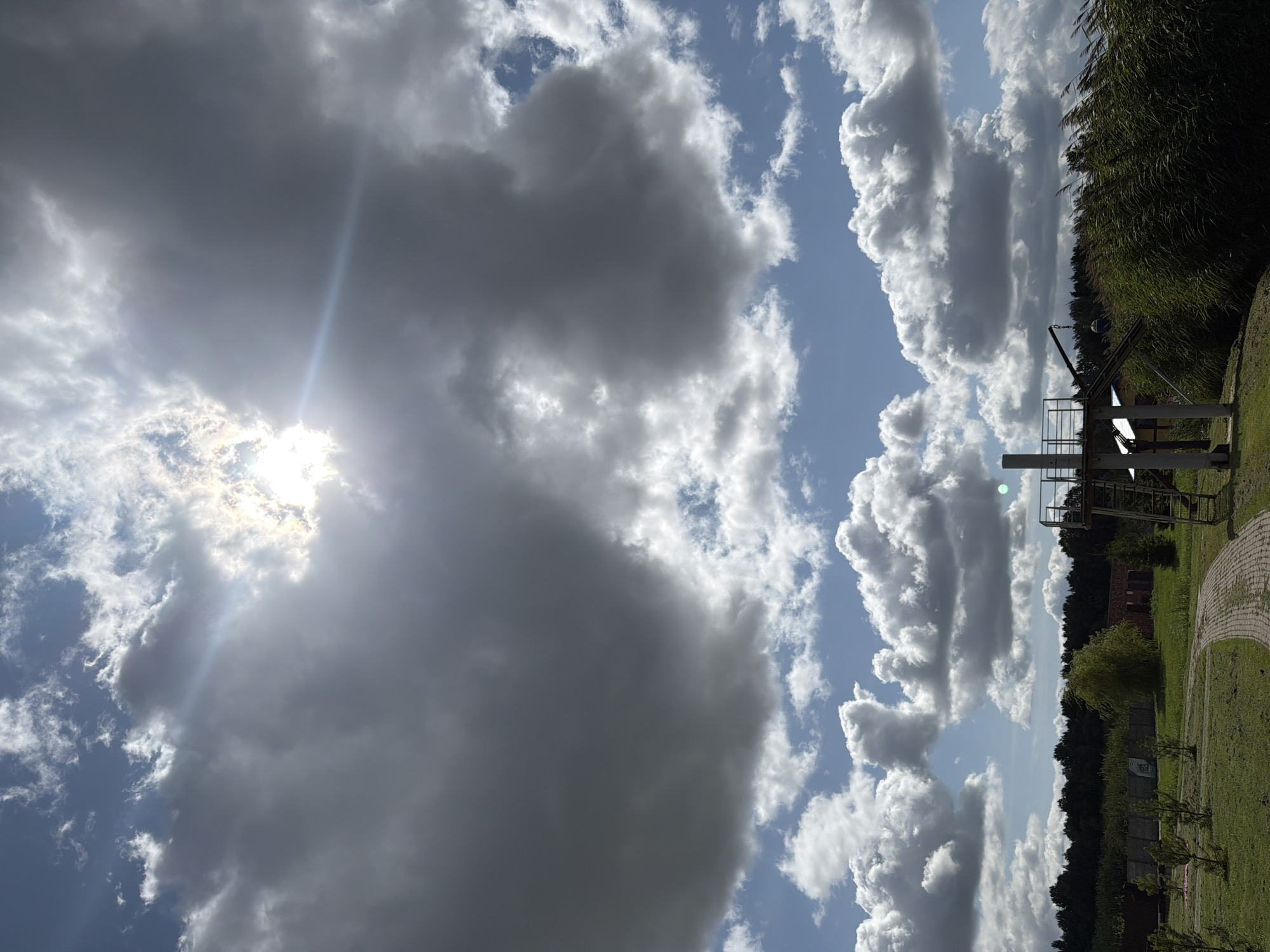 This image depicts a rural landscape under a dramatic sky. The upper portion of the frame is dominated by a mix of dark, heavy clouds and brighter, scattered cumulus clouds, with the sun peeking through, casting sunbeams. The lower half of the image shows a pathway made of red brick, winding through a grassy area. To the right of the path, tall, green reeds sway in the wind. In the background, a dense line of dark green trees forms a horizon, and a structure with a metal platform and stairs stands on the right side. There are no people visible in the image. The scene is set in Konstantinovka, Russia, and appears to be a tranquil, natural setting with some man-made elements.