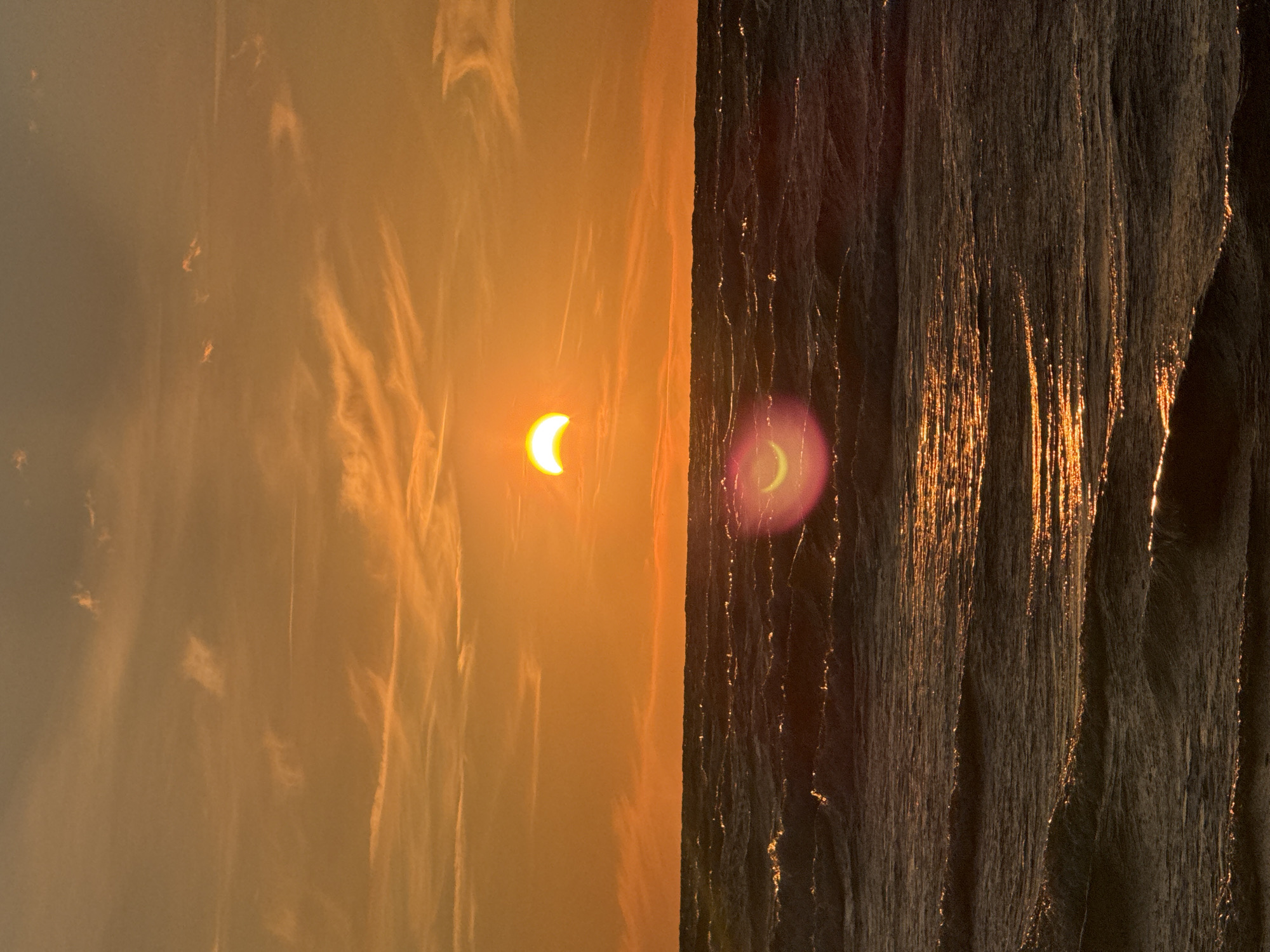The image shows a partial solar eclipse occurring over the sea during sunset. The sun, partially obscured by the moon, appears as a bright crescent in the upper portion of the frame. The sky is filled with orange and yellow hues, with wispy clouds streaking across it. The sea below is dark and choppy, with waves reflecting the warm light of the setting sun. A lens flare is visible in the lower half of the image, creating a soft, colorful glow. The scene appears to be captured from a beach or a viewpoint overlooking the sea, likely in Jūrmala, Latvia, given the characteristic Baltic Sea coastline and the typical sunset colors observed there. There are no people or specific structures visible in the frame that would definitively pinpoint a location within Jūrmala. The focus is on the natural phenomenon of the eclipse and the atmospheric conditions of the sunset.