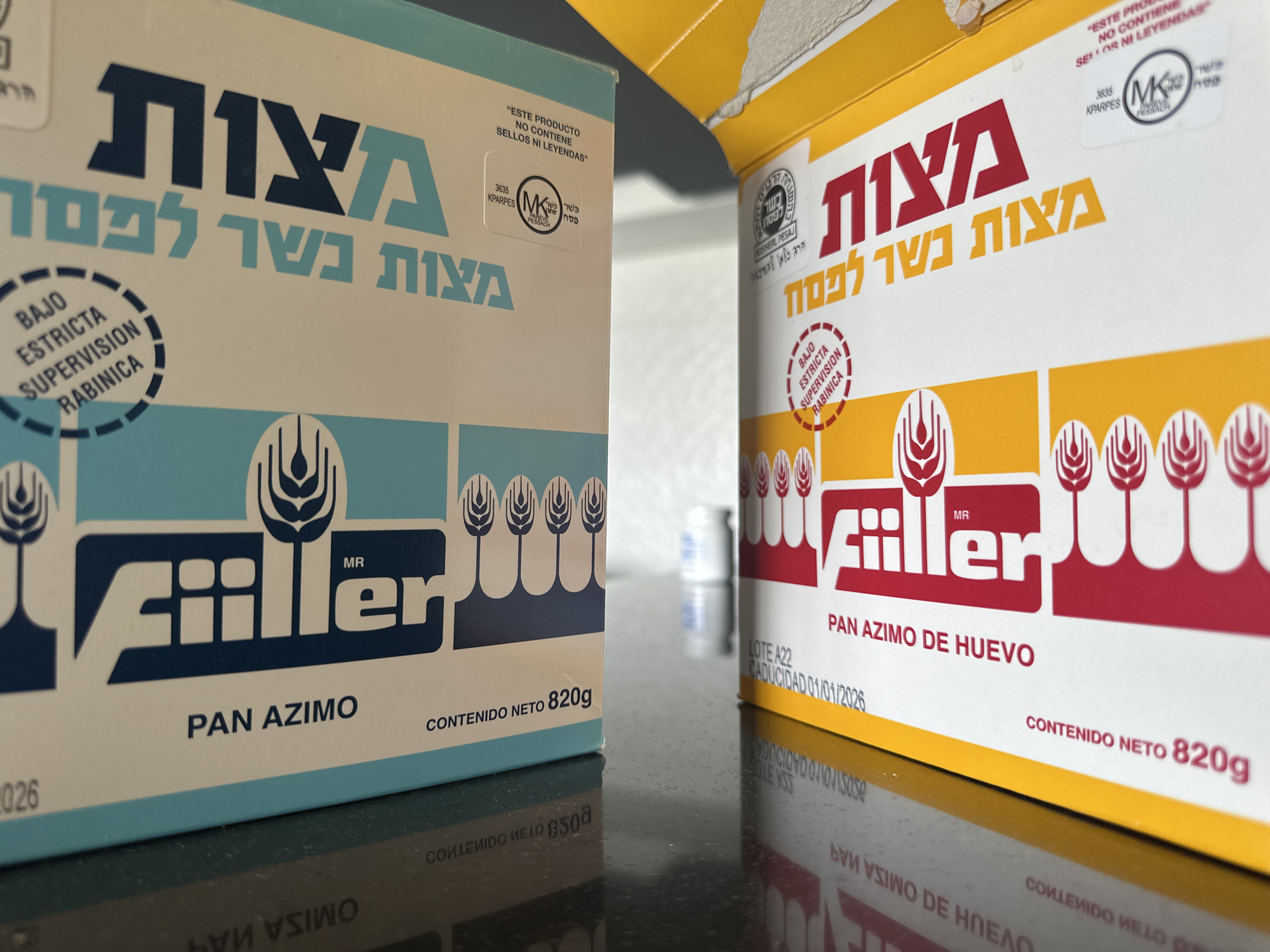 Two boxes of Mr. Filler brand "Pan Azimo" (unleavened bread) are situated on a dark surface. The box on the left has a blue and white color scheme, while the one on the right is yellow and white. Both boxes feature Hebrew lettering at the top, followed by the text "Mitzvot Kosher Le Pesach" (Mitzvot Kosher for Passover). The packaging includes a "Bajo Estricta Supervision Rabínica" (Under Strict Rabbinical Supervision) seal and a kosher certification (MK Pareve Pessach). Below the Mr. Filler logo, the blue box reads "Pan Azimo," while the yellow box specifies "Pan Azimo de Huevo" (Egg Unleavened Bread). The net weight of each package is stated as 820g. The yellow box also indicates a "Caducidad" (expiration date) of 01/01/2026 and mentions "LOTE A22" (Batch A22). The labels on both boxes state, "Este producto no contiene sellos ni leyendas" ("This product does not contain seals or legends"). The environment suggests a kitchen setting due to the reflective countertop. Huixquilucan de Degollado, Mexico is not specified.