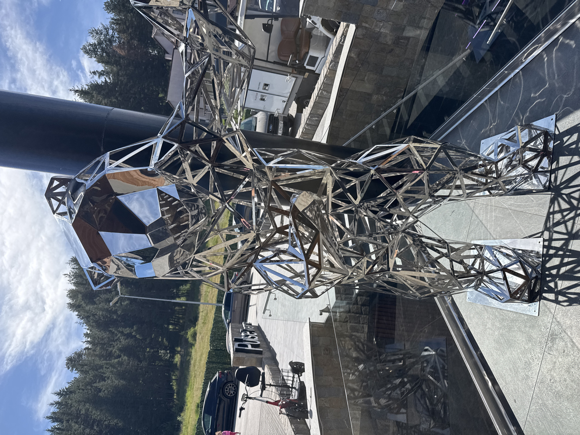 A polished metal sculpture of a bear is displayed outdoors on a stone surface. The sculpture is constructed of interconnected geometric shapes. In the background, there are trees, a building, and a blue sky with white clouds. The sculpture is near a building marked with the word "Pulse." The overall scene appears to be in an urban or suburban environment.