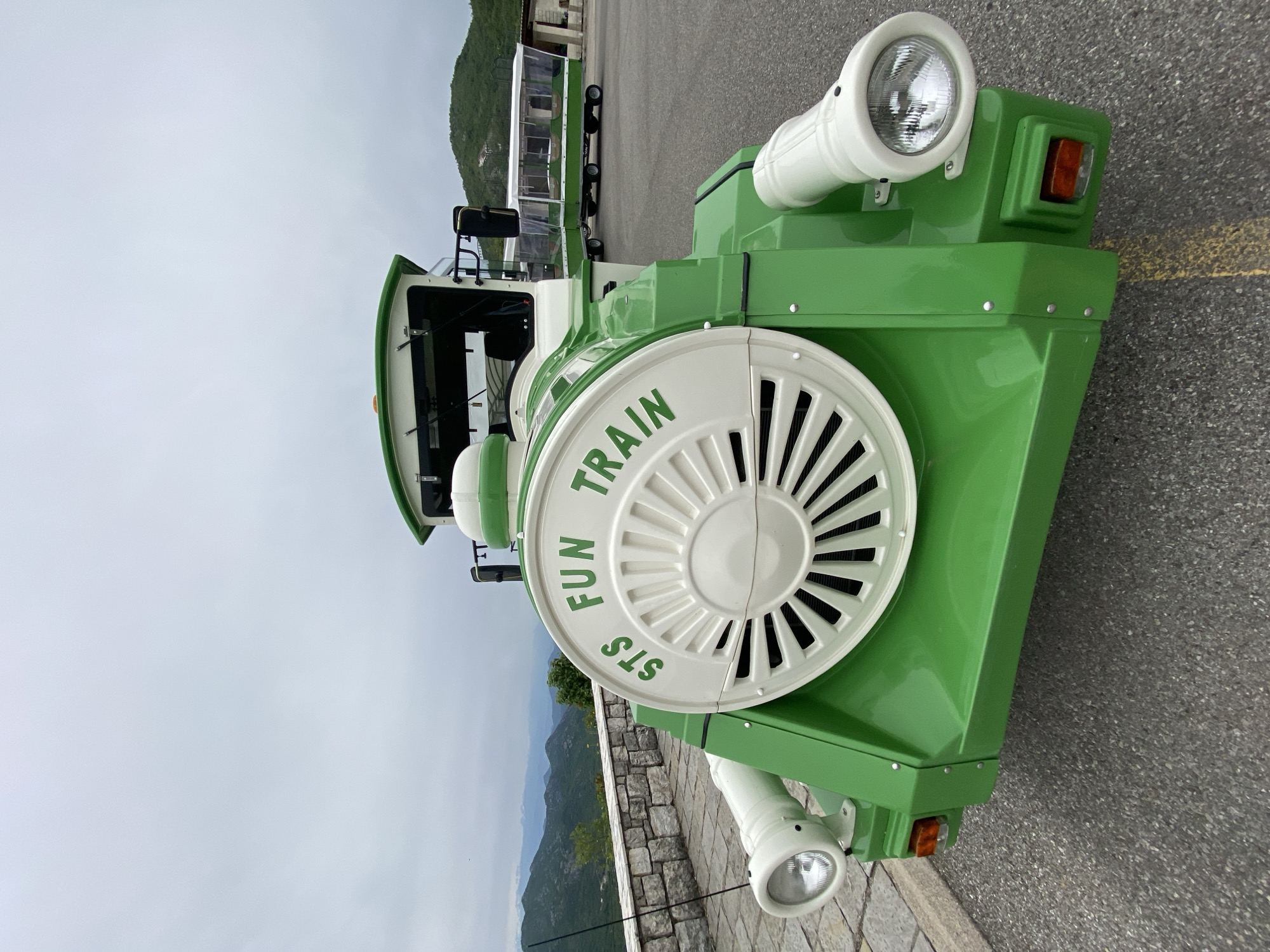 The image shows a "Fun Train" in Lipa Dobrska, Montenegro. The train is predominantly green and white. It has large, decorative white wheels with the words "Fun Train STS" printed on them in green. The train features white headlights and an orange turn signal. In the background, there is a mountain range and a cloudy sky. The train is parked on a grey paved surface with a low stone wall visible in the mid-ground. Additional train cars are visible behind the engine.