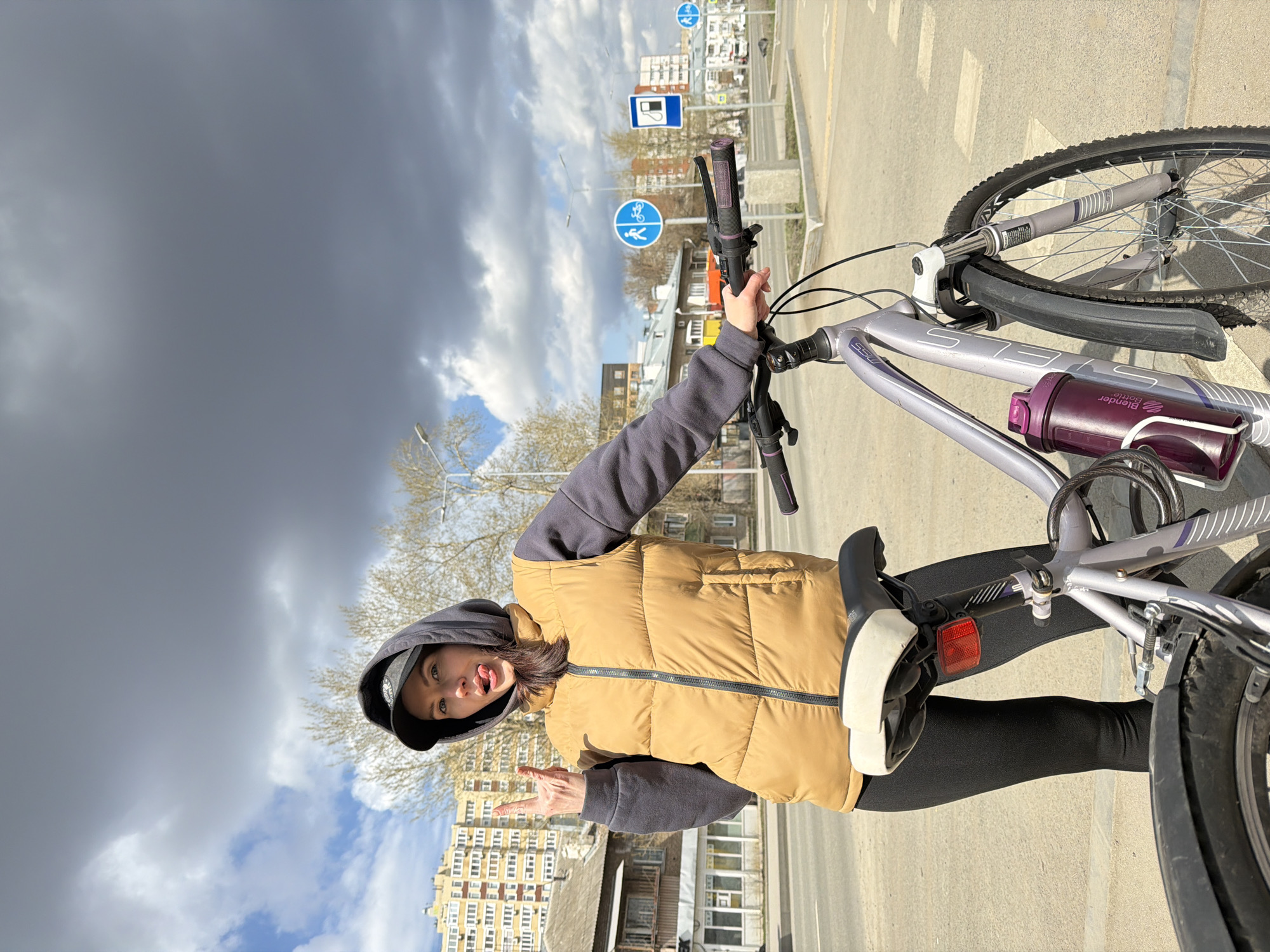 The image, taken in Perm, Russia, captures a young woman posing playfully next to her bicycle on an urban street during the daytime. The sky above presents a dramatic contrast, featuring large, dark grey clouds dominating the upper left, while patches of bright blue sky with white clouds are visible towards the right and bottom, suggesting dynamic weather conditions.
The woman is positioned to the left of her bicycle, wearing a dark hooded sweatshirt, a light brown/tan puffer vest with a black zipper, black leggings, and a black baseball cap. She holds the bicycle handlebars with her left hand while playfully sticking her tongue out and making a "rock on" hand gesture with her right hand, conveying a cheerful and confident mood.
Her bicycle is a silver/light grey model, with "STELS" visible on its frame. It features a purple "BlenderBottle" brand water bottle attached to the frame and a cable lock wrapped around the top tube. A red reflector is visible beneath the bike's seat.
In the background, an urban environment unfolds with multi-story apartment buildings, an asphalt road with white markings, and leafless trees, characteristic of an early spring setting. Several road signs are visible; two prominent blue circular signs feature white pictograms: one indicating a shared pedestrian and cyclist path, and another depicting a gas pump symbol, suggesting a nearby gas station. A lamppost is also part of the streetscape. The overall scene is a candid snapshot of a casual moment in a city.