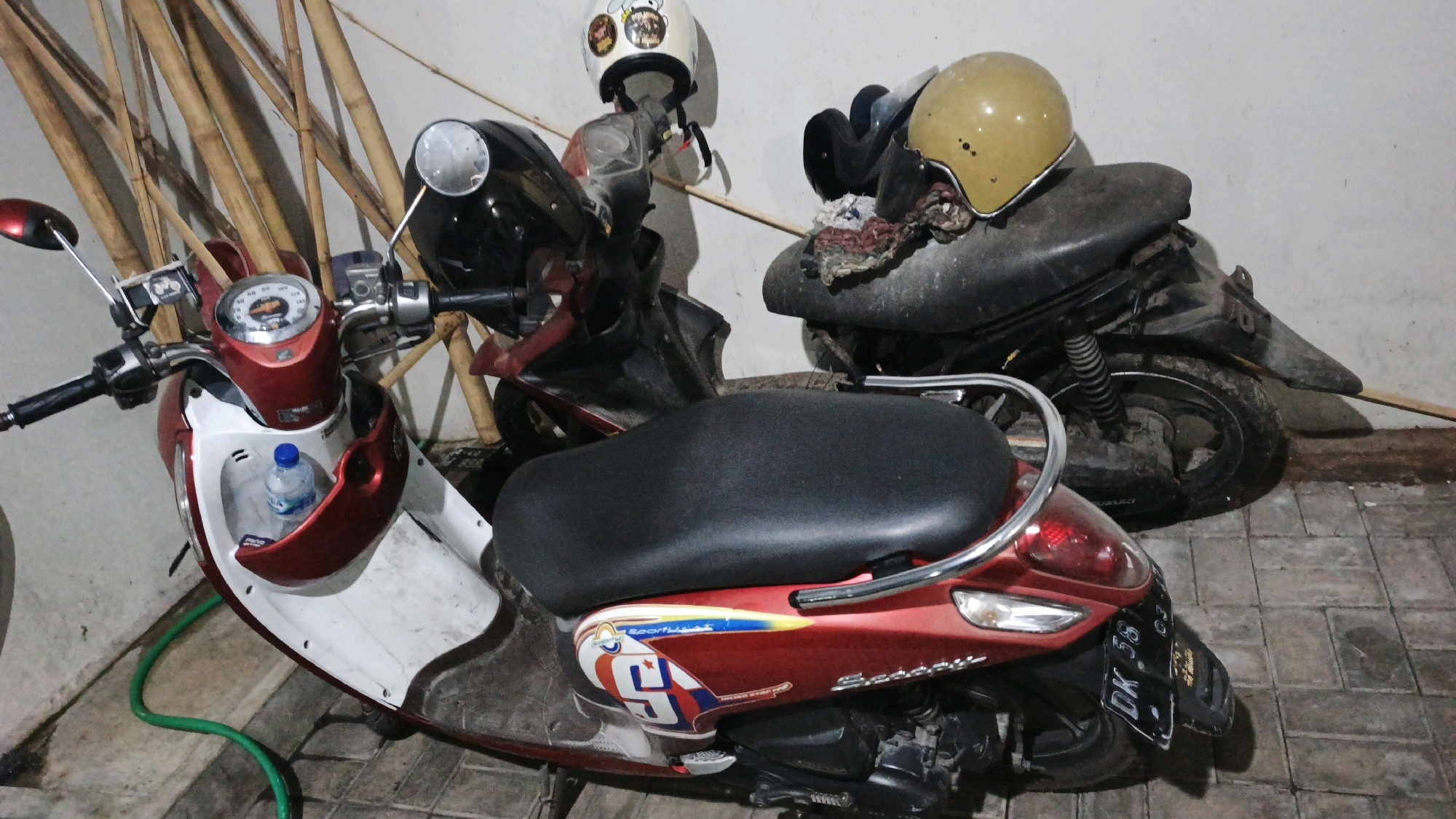 The image displays two scooters parked in close proximity against a wall. The scooter in the foreground is red and white, with a black seat and chrome accents. It has a water bottle in the front storage compartment and a green hose coiled on the ground near its front wheel. A license plate reading "DK 583" is visible on the rear of this scooter. The second scooter, partially obscured, is darker in color and has a yellow helmet resting on its seat. Both scooters appear to be parked on a paved surface. The background includes bamboo poles and a white wall. The scene suggests a residential or storage area, possibly a garage or a backyard, within the city of Denpasar, Indonesia.