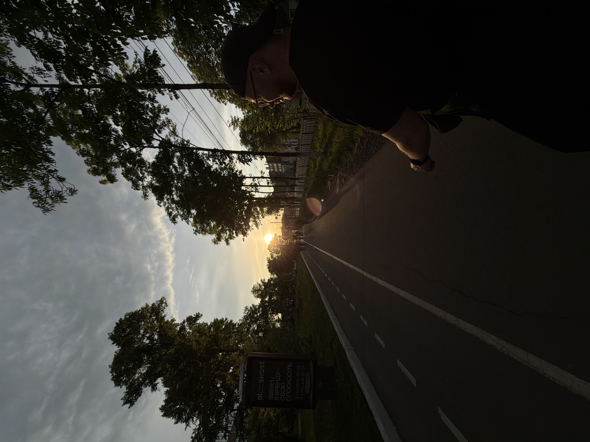 An outdoor scene in Birobidzhan, Russia, is captured during either sunrise or sunset. A paved path or road, marked with white dashed lines, extends into the distance, flanked by trees and grassy verges. The low sun is prominent towards the end of the path, emitting a warm glow and causing a noticeable lens flare. The sky above is partly cloudy.
On the right side of the frame, a person, seen in partial silhouette, is positioned from the chest up, appearing to look forward or down the path. They are wearing a dark t-shirt, a cap, and glasses. A smartwatch is visible on their left wrist.
On the left side of the path, a rectangular sign stands among the trees. The visible Russian text on the sign reads: "ploskost", "Забери", "Варенду-", "Свою", "плоскость", followed by two phone numbers: "+7 (4212) 52-90-90" and "+7 (914) 428-07-07". The overall ambiance suggests a calm and contemplative moment during twilight.