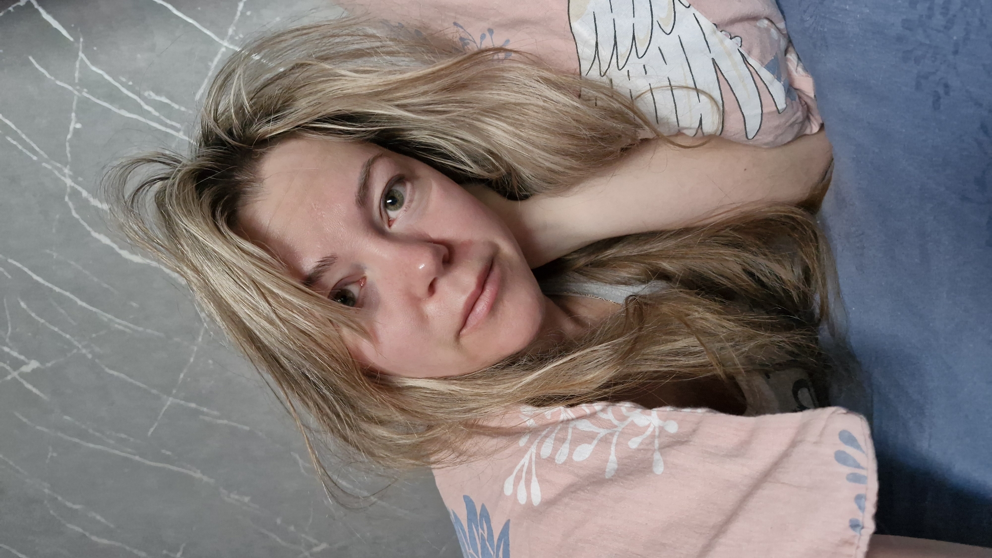 A woman with blonde hair is lying down. She is resting her head on a pillow with a pink and white pattern. She is looking directly at the camera with a neutral expression. The environment appears to be indoors, possibly a bedroom. The background includes a gray surface with a marble-like pattern and a blue surface. There is no indication of the location being Otchor, Russia.