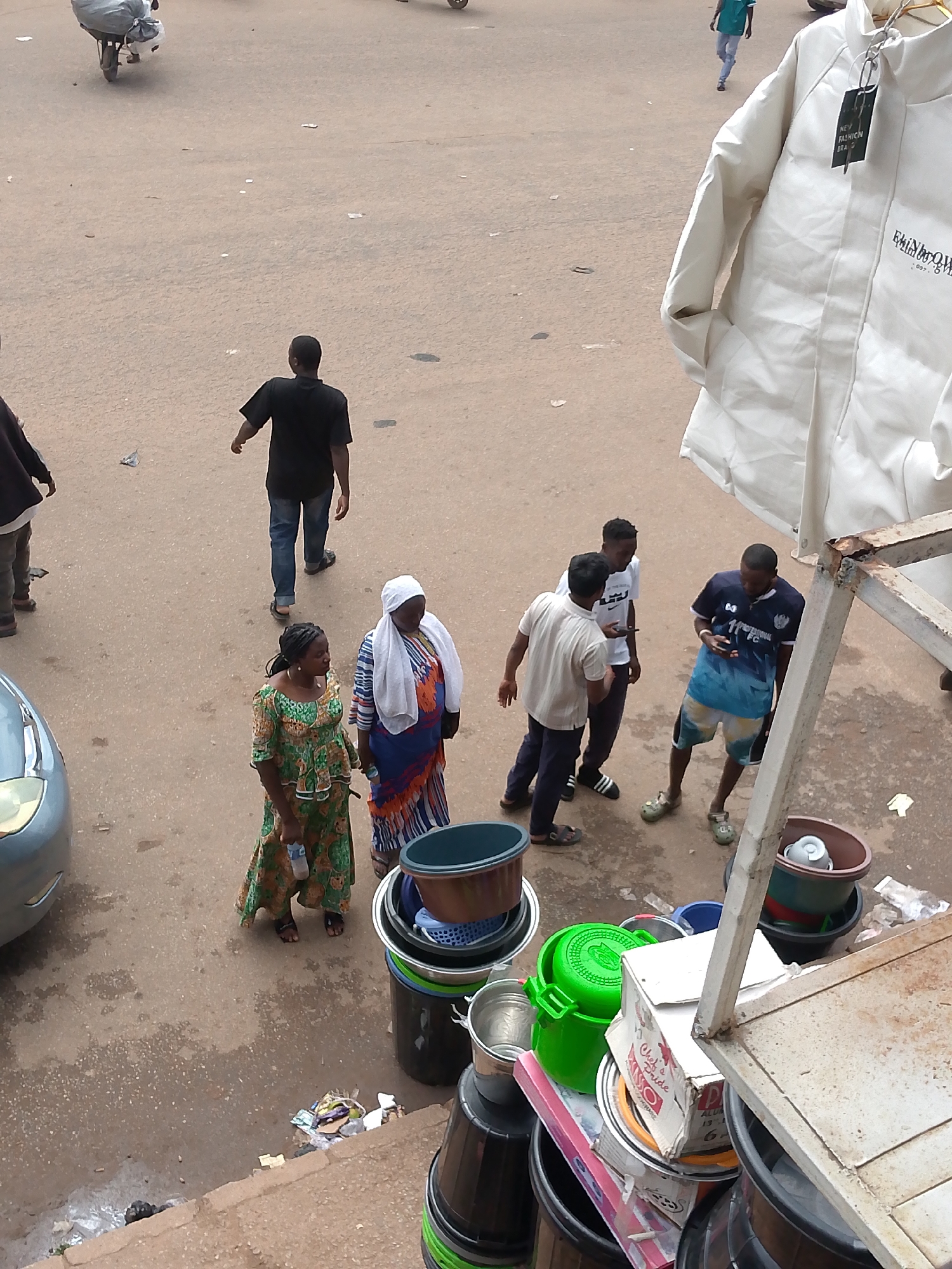 The image depicts a street scene in Bukuru, Nigeria, featuring several individuals and various objects. In the foreground, to the right, is a white jacket hanging on a metal structure. Several people are scattered across the dusty, paved street. Two women, one in a green and orange patterned dress and the other in a blue and orange patterned outfit with a white headscarf, stand together. Nearby, a man in a black t-shirt and blue jeans walks away from the camera. Further to the right, three young men are gathered, two of whom appear to be in conversation. One man wears a dark blue t-shirt with a blue and white patterned design on his shorts, while another wears a white t-shirt and dark pants. The third man, in a light-colored t-shirt and dark pants, is partially obscured. Various items, including buckets, containers, and a car on the left side of the image, are visible on the ground. A wheelbarrow loaded with a large white bundle is also present in the upper left portion of the frame. The environment suggests an outdoor market or a busy street.