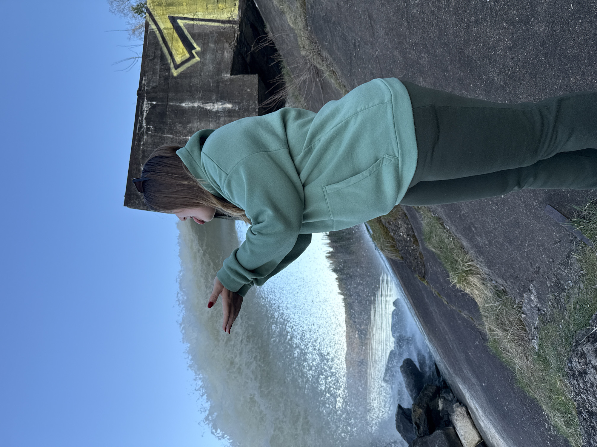 A woman in a light green hoodie and dark green pants stands at the edge of a concrete structure, reaching out her hands towards a cascading waterfall. The waterfall flows into a rocky area. Behind the woman is a concrete wall with some sort of structure and graffiti. The sky is blue. The location is Ol'khovskoye, Russia.