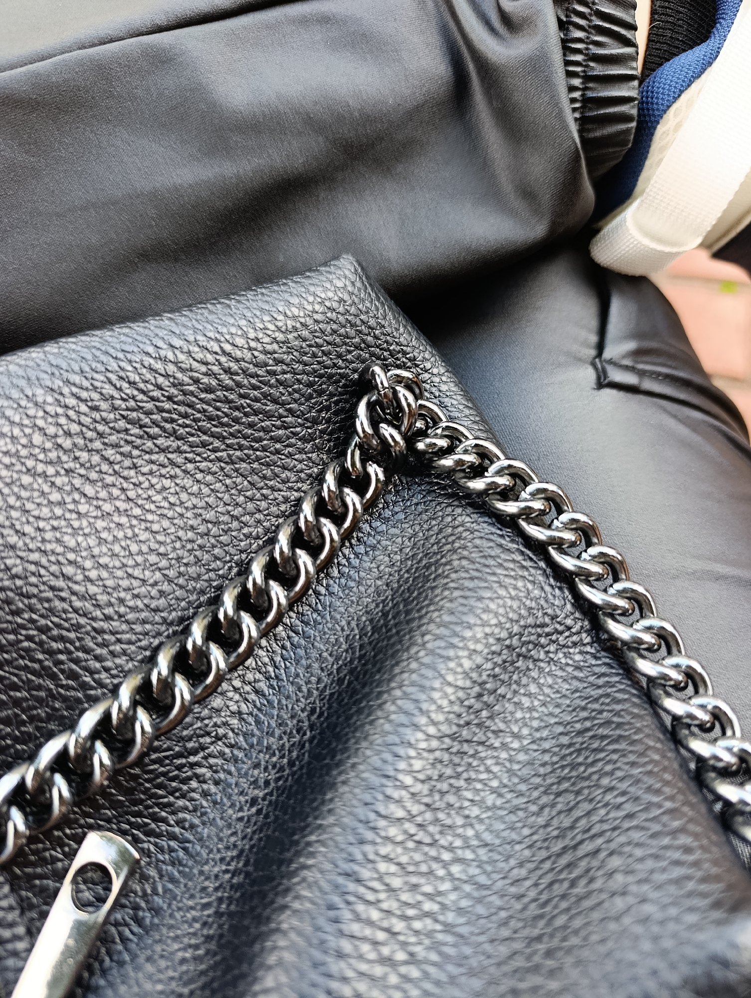 A close-up, eye-level shot shows a textured black leather purse with a metal chain strap. The purse is resting on dark, smooth fabric, likely clothing. In the upper right corner, a portion of a white sneaker with blue accents is visible. The background is softly blurred, suggesting an outdoor setting. The lighting is natural, casting subtle shadows. This image was captured in Kyiv, Ukraine.