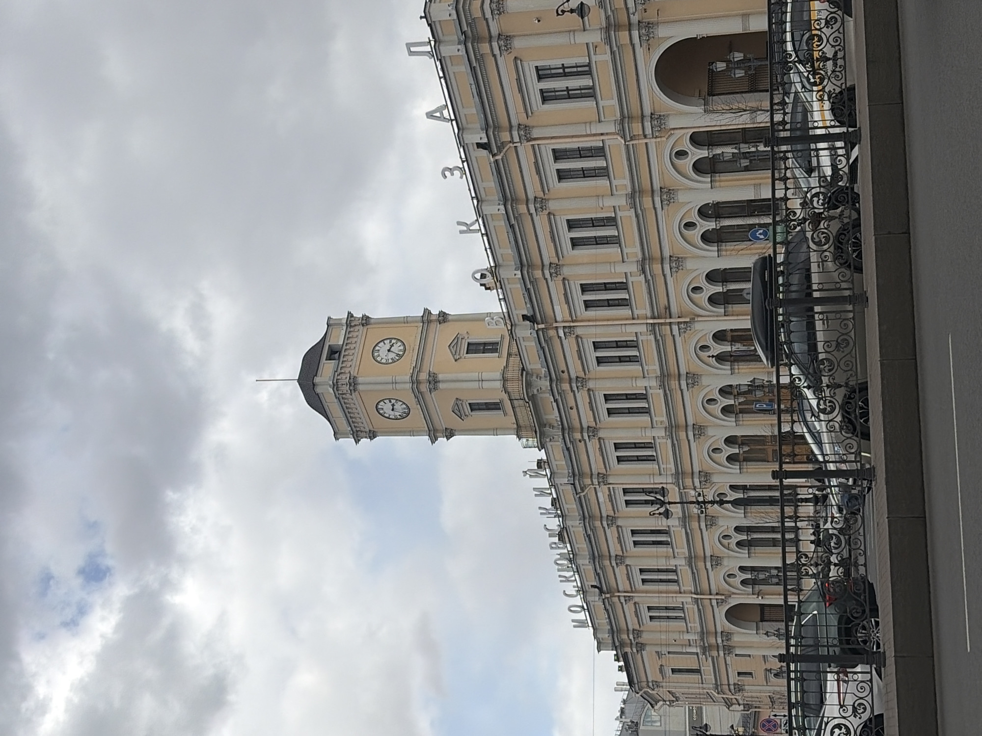 The image, when rotated correctly, depicts a grand, multi-story building with a prominent clock tower under an overcast sky. The architecture is classical or neo-classical, characterized by a light yellow or beige facade with white/grey trim, decorative pilasters, cornices, and numerous arched windows and entrances. A clock face on the tower shows the time, approximately 2:15 or 3:15.
In the foreground, an ornate black wrought-iron railing separates the building from a street where several cars are parked, including a black sedan and a white van or bus partially visible. The sky is cloudy and grey, indicating an overcast daytime scene.
Large, illuminated letters are mounted on the roofline of the building. These letters spell out "МОСКОВСКИЙ ВОКЗАЛ" (Moskovsky Vokzal, meaning Moscow Railway Station) and "ПЛАЗА" (Plaza). This text, along with the distinctive architecture, identifies the location as Moskovsky Vokzal in Saint Petersburg, Russia. No specific activities or people are prominently featured, portraying a static urban landscape.