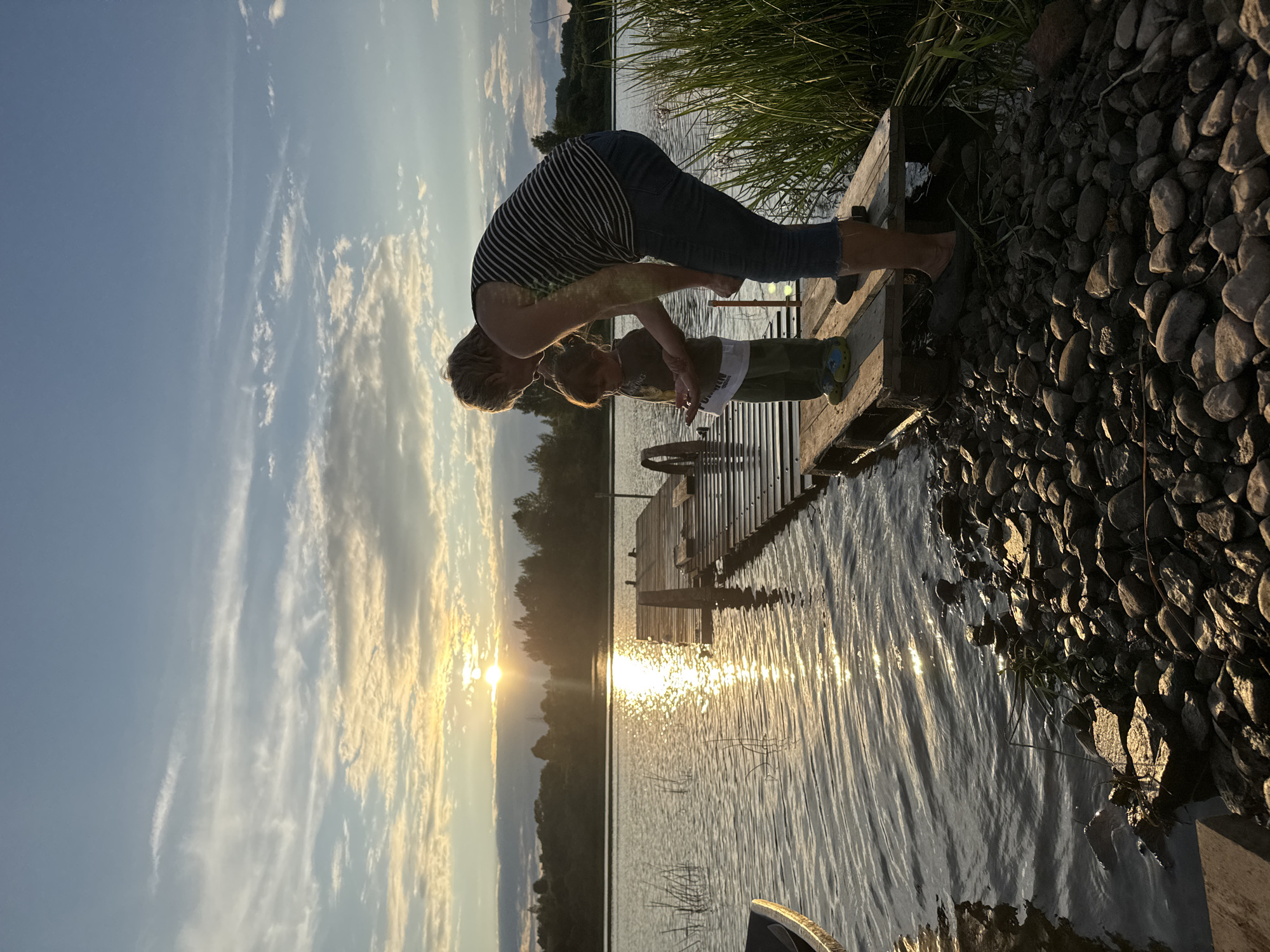 A woman and a young child stand on a wooden dock at the edge of a body of water, likely a lake or river, in Sitno, Russia. The sun is setting, casting a warm glow across the scene and reflecting on the water's surface. The sky is a mix of blue and white clouds, with the sun partially obscured by them. The dock is constructed from weathered wood and extends out over the water. The child, facing away from the camera, appears to be looking at something in the woman's hand. The woman is bent over, interacting with the child. The shoreline is lined with rocks and tall grasses. The overall atmosphere is peaceful and serene, with a hint of familial connection.