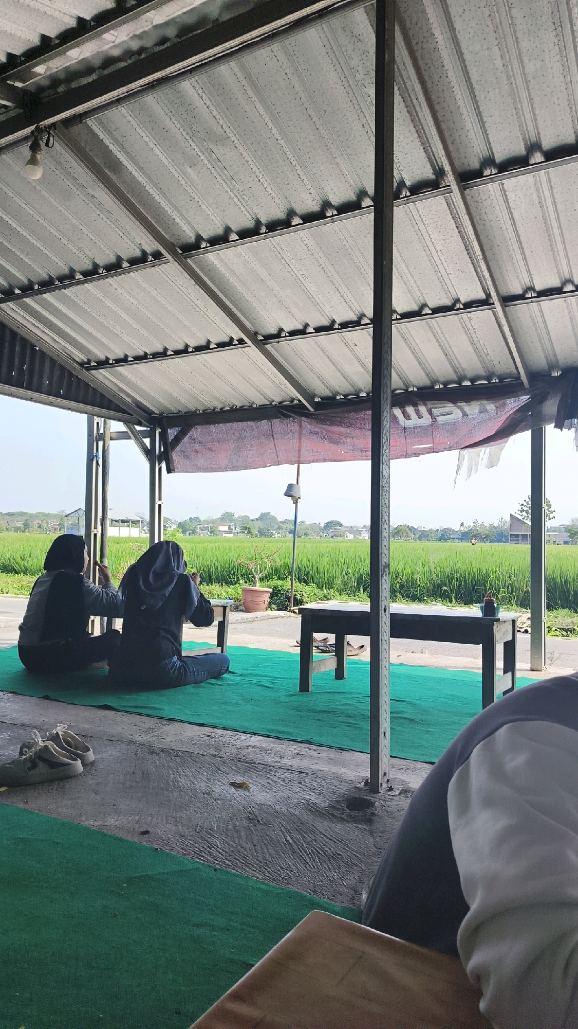 In Ngemplak, Indonesia, two individuals are seated on a green carpet under a metal corrugated roof. The person on the left wears a dark jacket and a hijab, while the person on the right wears a dark blue jacket and hijab. They are facing a lush green rice paddy field, with a line of trees and houses in the distance under a bright sky. A dark wooden table is positioned in front of them, with a small bottle of sauce on it. Another table is visible to the right. A pair of white sneakers are placed on the concrete floor to the left. The scene is an outdoor seating area overlooking agricultural land.