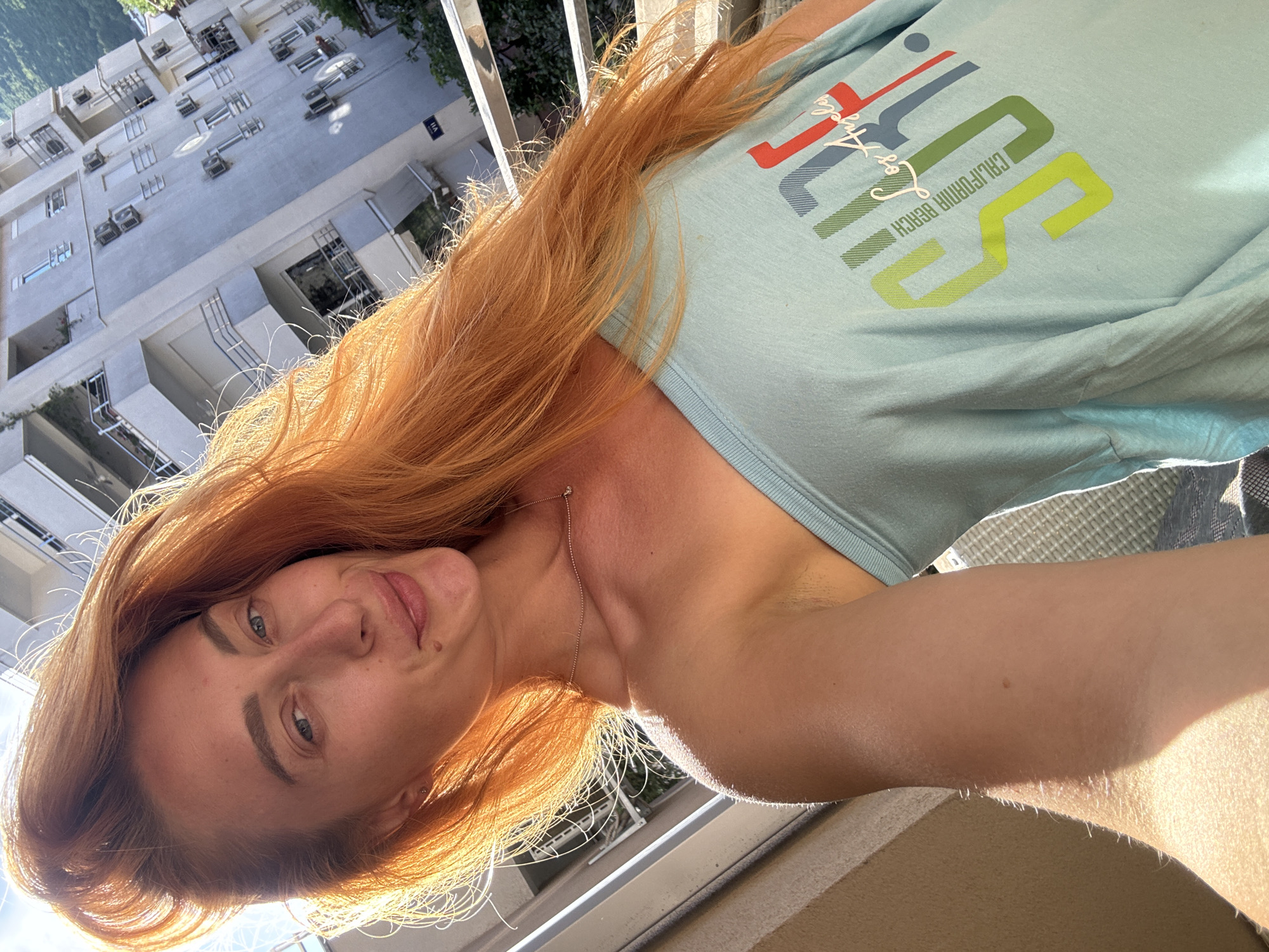 A woman with long, bright orange hair is taking a selfie on a balcony in Budva, Montenegro. She is wearing a light blue, off-the-shoulder t-shirt with a graphic design that appears to say "SURF CALIFORNIA BEACH Los Angeles." Sunlight is hitting her hair, creating a strong backlight. In the background, a multi-story apartment building with balconies and air conditioning units is visible, along with trees and a hint of mountains. The woman has fair skin and is smiling slightly. The overall scene suggests a bright, sunny day.