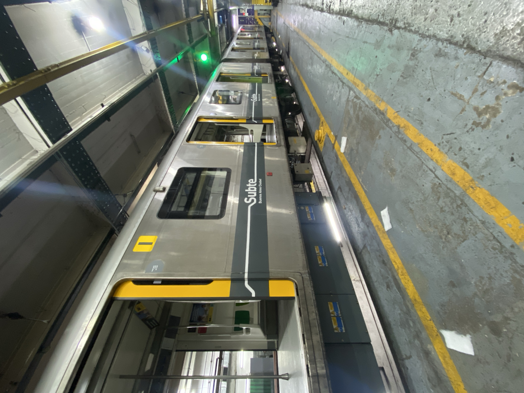 The image shows a silver subway train of the Subte (Subterraneos de Buenos Aires) in Buenos Aires, Argentina. The train car is mostly silver with yellow accents around the doors and a dark gray vertical stripe with the word "Subte" written on it. It is parked inside what appears to be a maintenance depot. The floor alongside the train has yellow lane markings. The train doors are open, revealing the interior. The ceiling of the depot is visible above the train. There are no people visible in the frame.