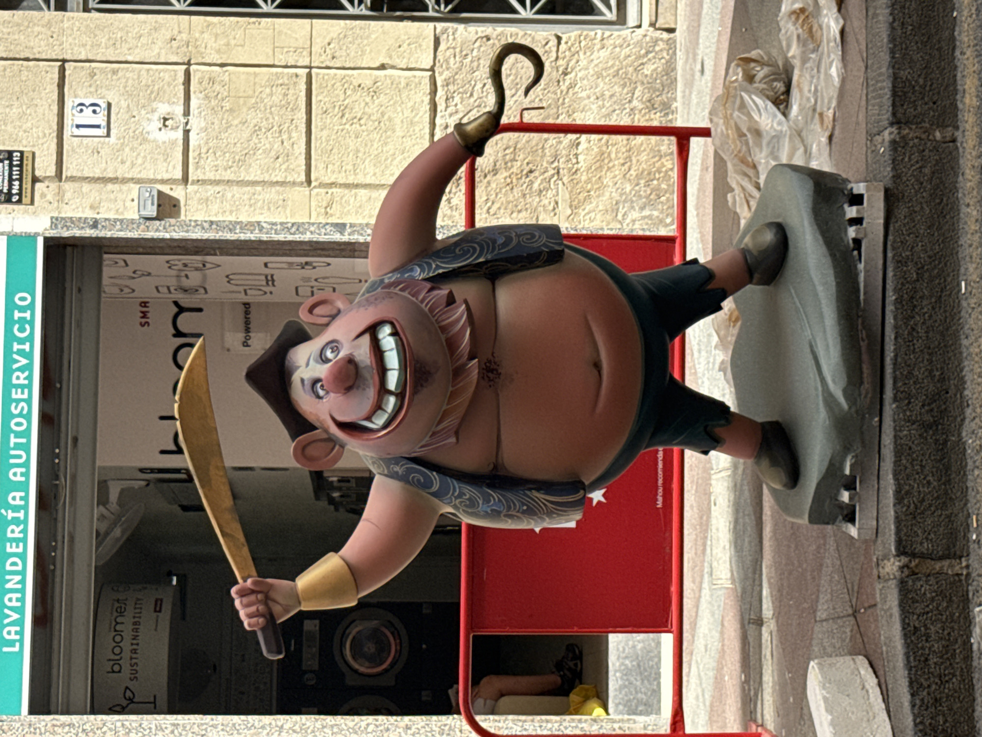 A statue of a caricature of a rotund, red-faced pirate with a large grin stands on a grey base on a sidewalk. The pirate wears a dark blue vest with gold trim, a brown hat, and dark green pants. He holds a curved, golden sword in his right hand and has a hook for a left hand. Behind the statue is a red barrier. In the background, a storefront displays a sign that reads "LAVANDERÍA AUTOSERVICIO" and "bloomeet SUSTAINABILITY". There are also washing machines visible inside the storefront. A building with a numbered sign "13" is visible to the right of the storefront. The ground is paved with concrete and asphalt.