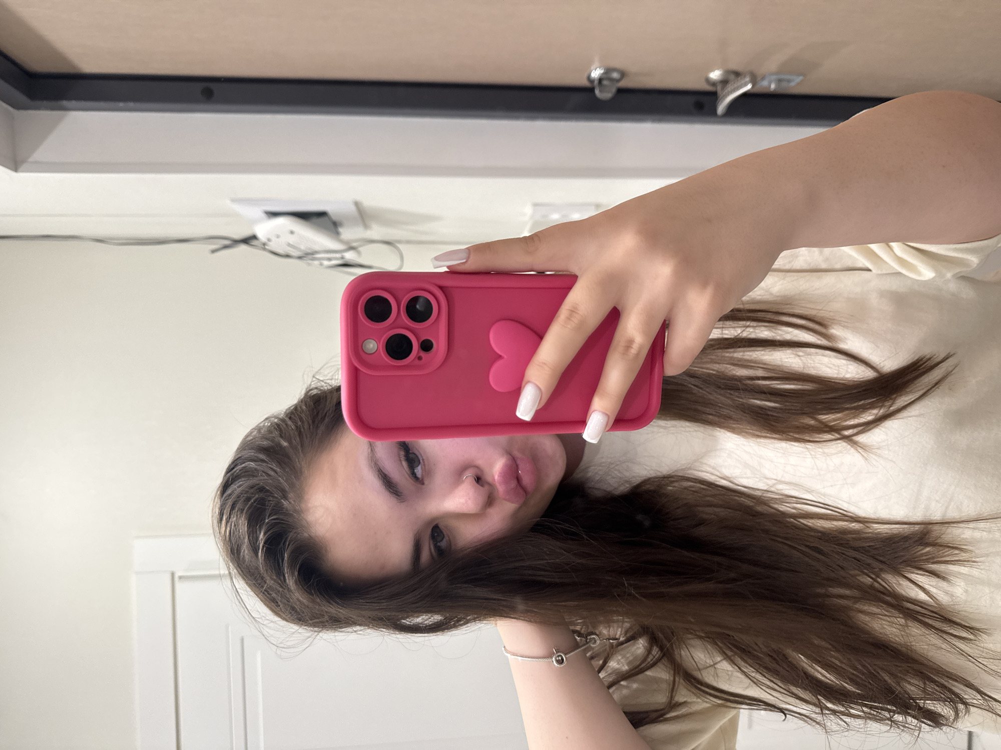 A young woman is taking a selfie indoors. She is holding a pink iPhone with a heart-shaped case. She has long brown hair, fair skin, and is wearing a light-colored shirt. She is making a kissing face. The background shows a white wall with electrical outlets and a door. The location is presumed to be Murino, Russia, but there is no direct evidence to confirm this from the image.