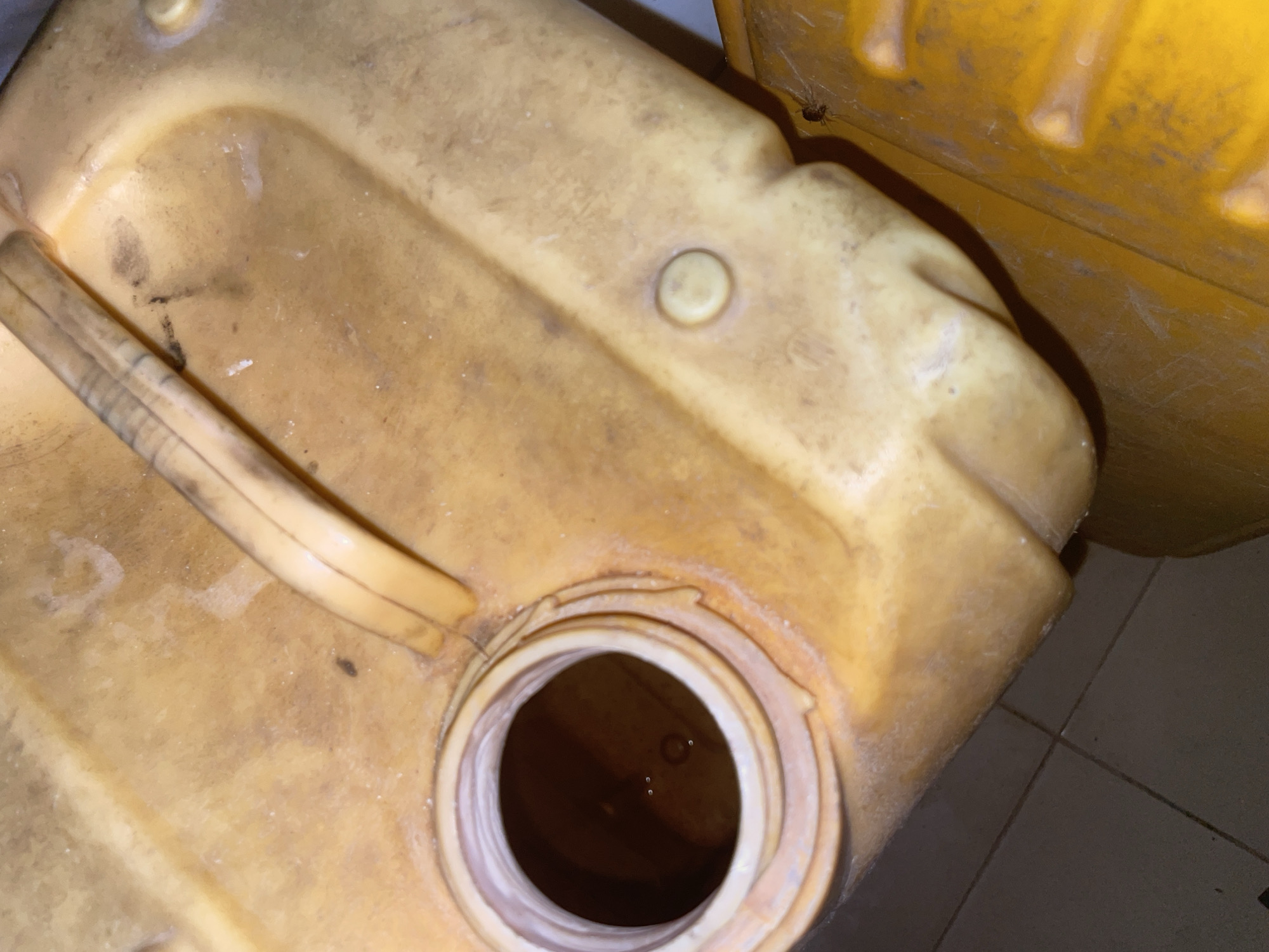 A close-up, high-angle shot shows the opening of a large, yellow plastic jerrycan filled with a dark liquid. The jerrycan has a textured handle and a circular opening with a white rim. The exterior of the jerrycan is dusty and appears stained in places. Below and to the right of the jerrycan, another yellow plastic object is visible, possibly another jerrycan, with a ribbed surface. The floor beneath is tiled in a dark, square pattern. A small spider is visible on the ribbed surface of the lower yellow object. The scene suggests an outdoor or storage area in Jalingo, Nigeria, given the presence of the jerrycan, commonly used for water or other liquids in such regions, and the tiled flooring often found in residential or commercial spaces. There are no people or explicit actions depicted.