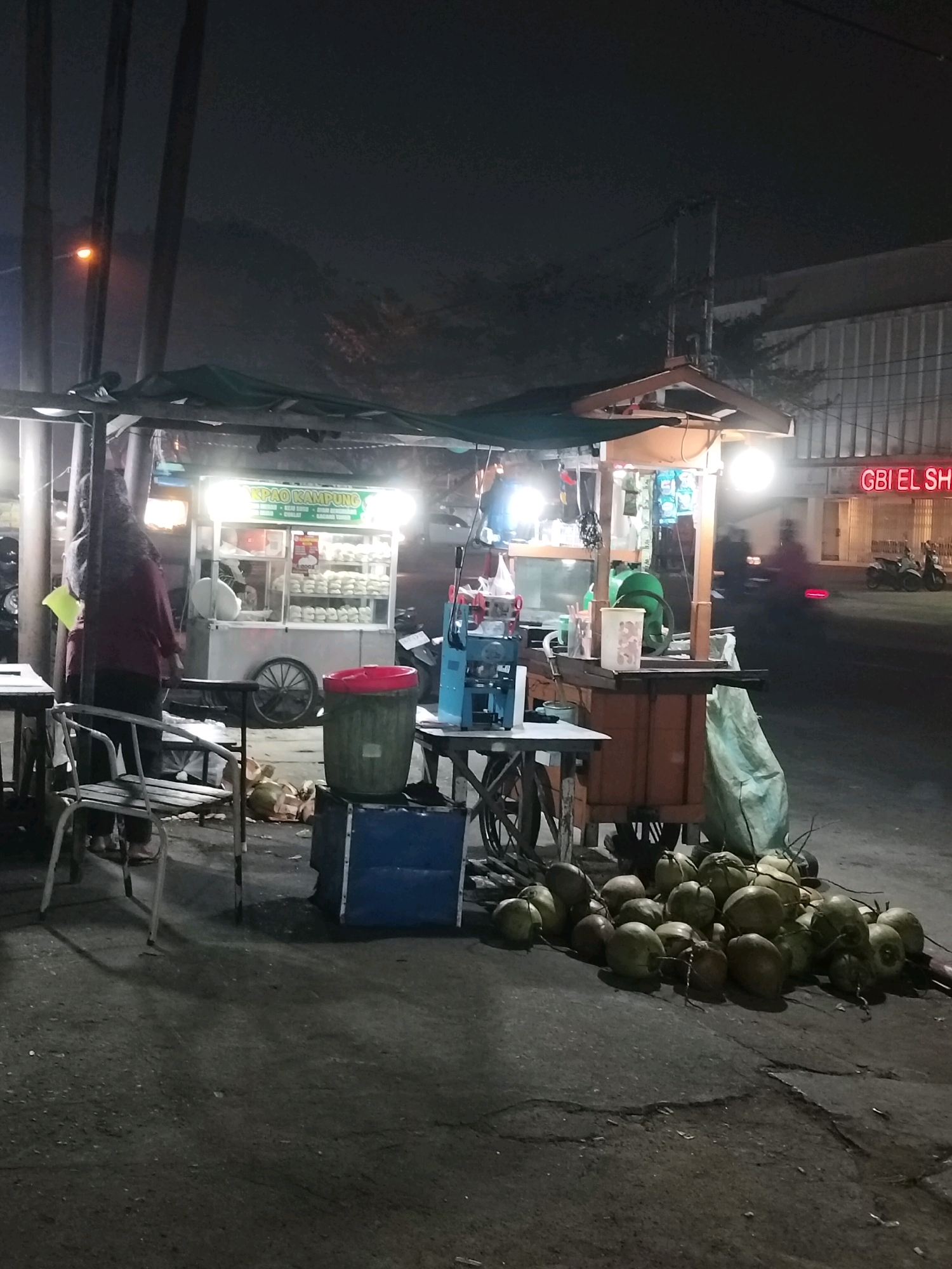 This is a nighttime photograph of a street food stall in Pontianak, Indonesia. The stall features a cart selling steamed buns, indicated by the sign "KPAO KAMPUNG." There are several coconuts piled on the ground in front of the stall. To the right of the stall, a building with neon signage that reads "GBI EL SH" is visible. The street is paved and appears to be wet, suggesting recent rain. The overall atmosphere is that of a typical Indonesian night market or roadside food vendor.