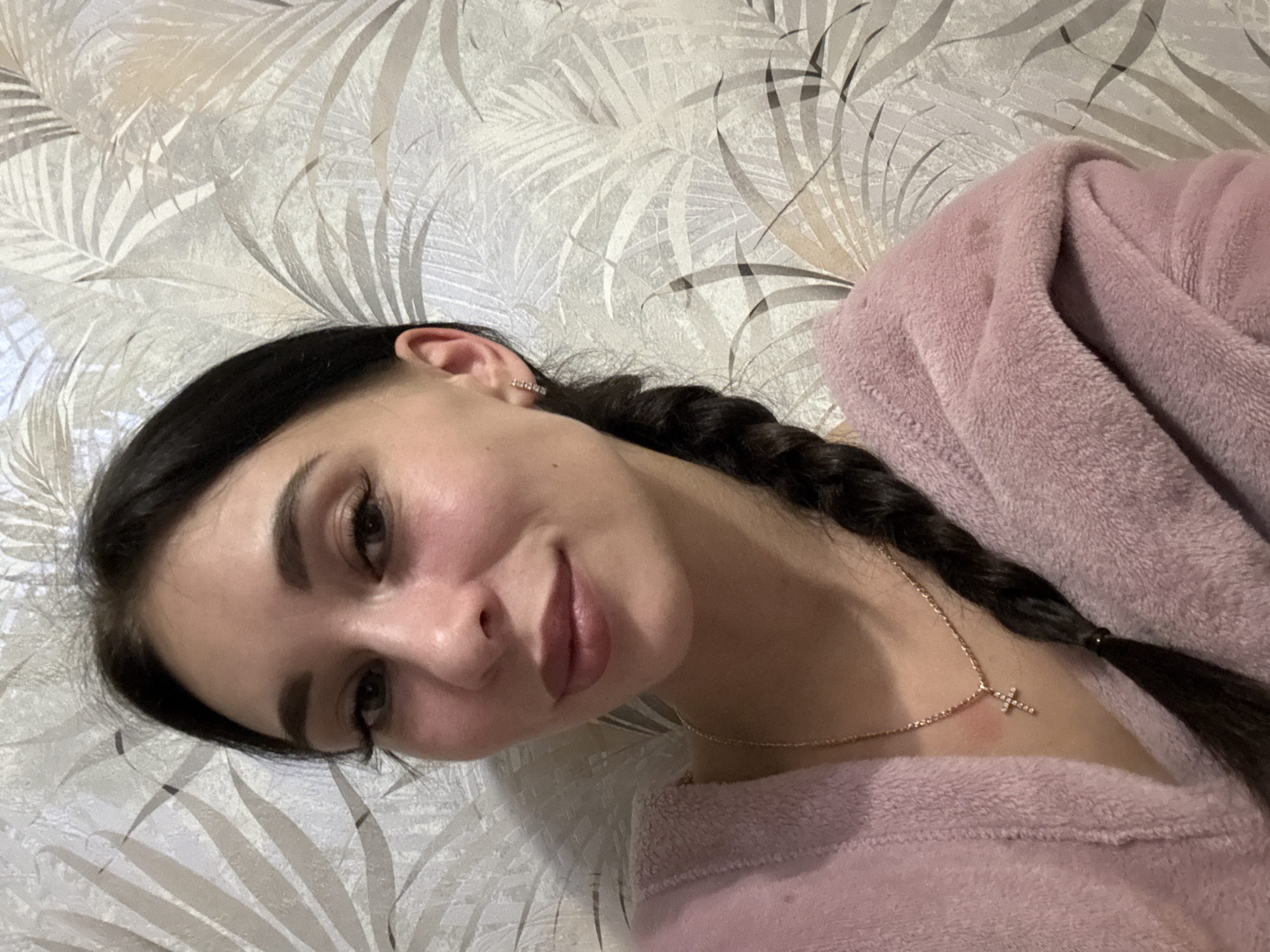 A young woman is pictured indoors, lying down. She has dark hair styled in a braid, is wearing a pink robe, and a delicate gold necklace with a cross pendant. The background features a wallpaper pattern of stylized palm leaves in muted shades of gray and white. The photograph appears to be taken at close range, likely from an overhead perspective. There is no information to suggest this image was taken in Chaikovsky, Russia.