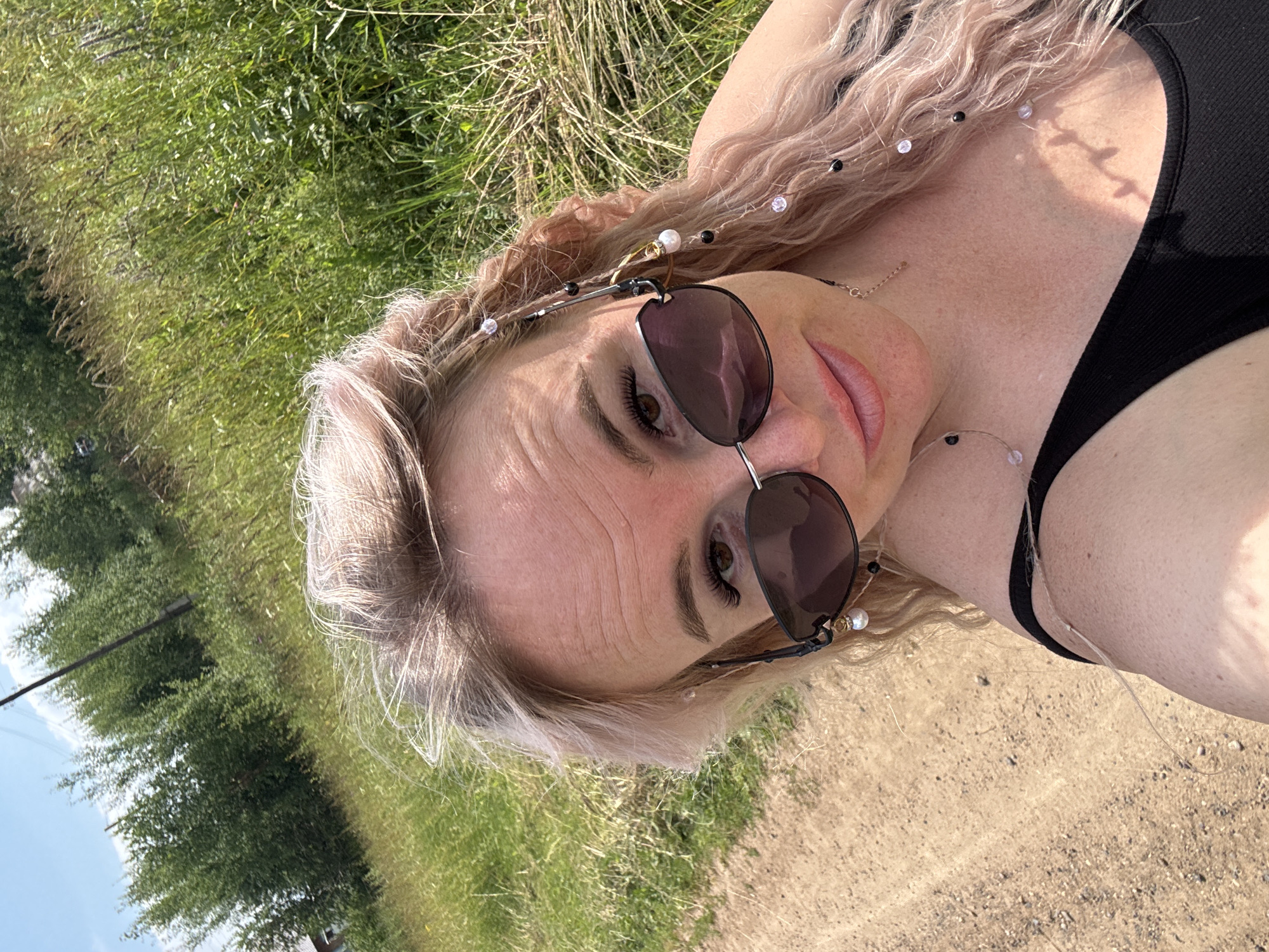 The image shows a close-up, selfie-style photograph of a woman outdoors, likely in the village of Pozdyshki, Russia, given the context. The woman has light-colored, wavy hair and is wearing dark sunglasses and a black tank top. Her hair is adorned with a decorative chain made of small black and white beads, attached to her sunglasses. The background consists of lush green grass, dry grass, and a dirt path. In the distance, trees and a utility pole are visible, suggesting a rural or semi-rural setting. The lighting indicates it is daytime, with bright sunlight casting shadows. The woman is looking towards the camera with a slight smile.