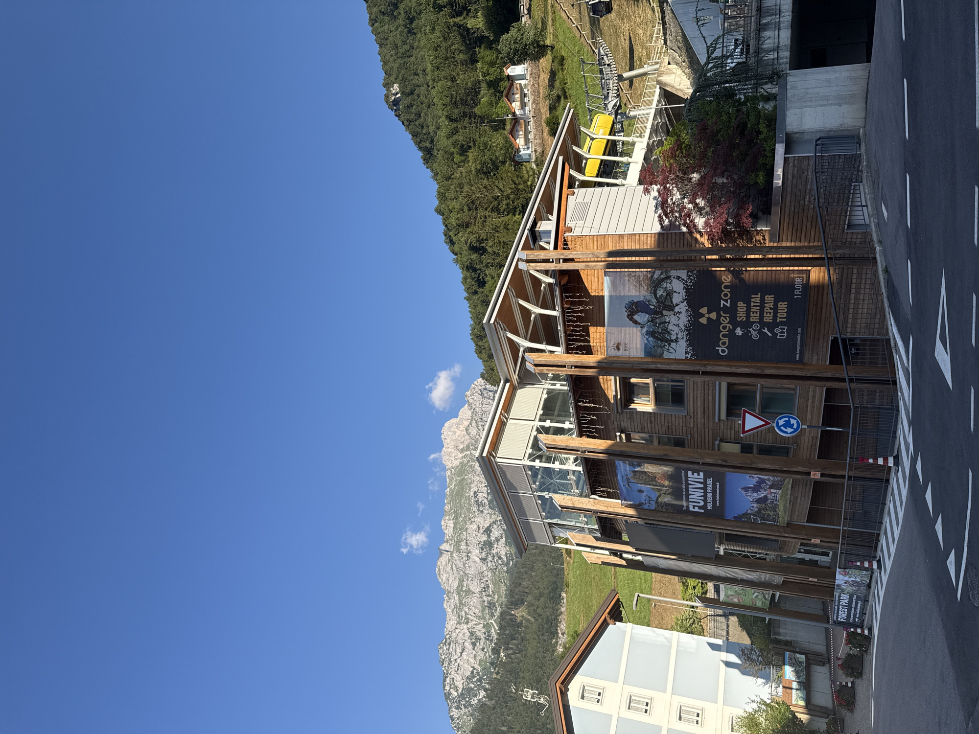 The image shows a sunlit, mountainous landscape in Molveno, Italy. In the foreground, a modern building with a wooden facade is visible. Large banners advertising "FUNIVIE" and "DANGER ZONE" are displayed on its side. The "DANGER ZONE" banner lists services such as "SHOP," "RENTAL," "REPAIR," and "TOUR." Further down, a sign for "FOREST PARK" is visible. A cable car system ascends a grassy slope covered with trees. The building is situated on a paved road with white directional arrows. In the background, a majestic mountain range with rocky peaks and forested slopes dominates the horizon under a clear blue sky. The overall scene depicts a tourist destination offering outdoor activities.