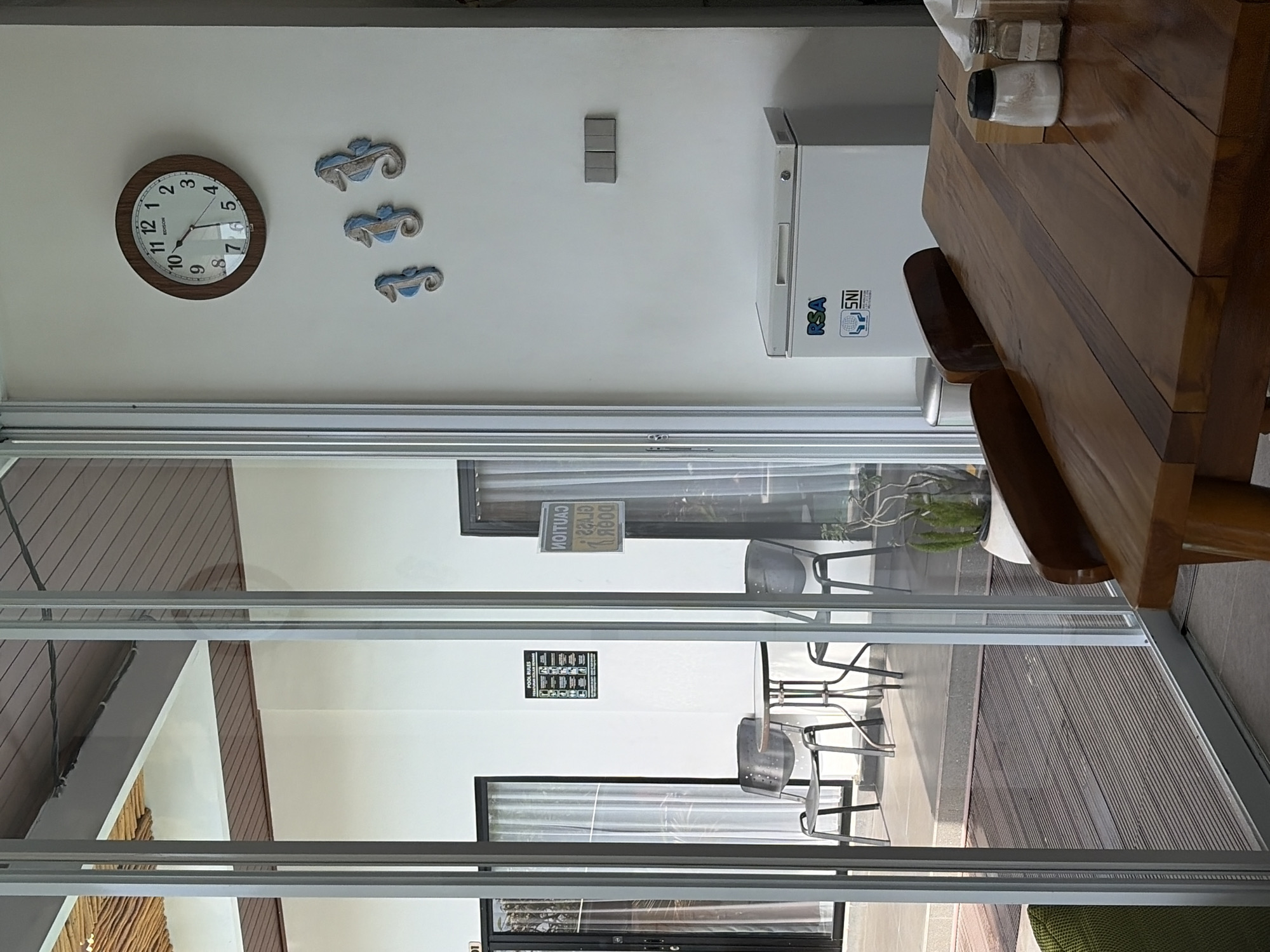 This is a full-frame, eye-level, indoor-outdoor shot of a room, possibly in Kubu, Indonesia, featuring a dining table in the foreground and a patio area visible through glass doors in the background. The room has white walls and a slatted wooden ceiling. A round, wooden-framed clock hangs on the wall, displaying the time as approximately 10:08. Below the clock are three decorative seahorse figures. To the right of the seahorses are three light switches. A white chest freezer with "RSA" and "SNI" stickers is positioned on the floor. In the background, through the glass doors, a patio with a small round table and two chairs is visible. There is also a sign on the wall that reads "POOL RULES". A wooden dining table occupies the bottom right portion of the frame, set with two spice shakers. The overall impression is one of a relaxed, possibly resort-like setting.
