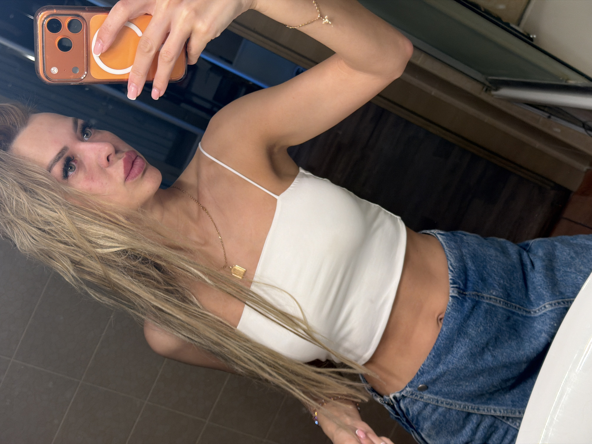A young woman with blonde hair and makeup is taking a mirror selfie in a bathroom. She is wearing a white strappy crop top and denim shorts. The background appears to be a tiled wall and a wooden vanity. The image was taken in Kyiv, Ukraine.