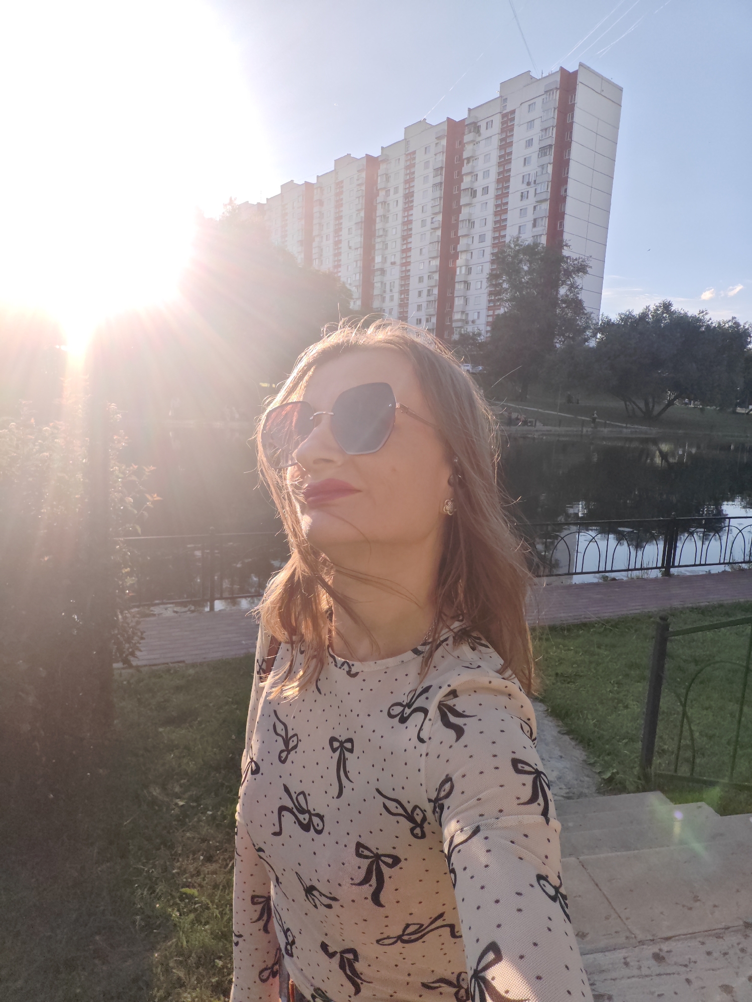A woman stands in front of a large apartment building with a body of water and trees in the background. The sun is shining brightly, creating lens flare in the image. The woman is wearing sunglasses and a patterned shirt. She is looking up and to the side, with her hair blowing in the wind. The apartment building is tall and white with red accents. The water is dark and still, reflecting the sky. There are trees with dark green foliage on the far bank of the water. The scene appears to be a park or recreational area within a city.