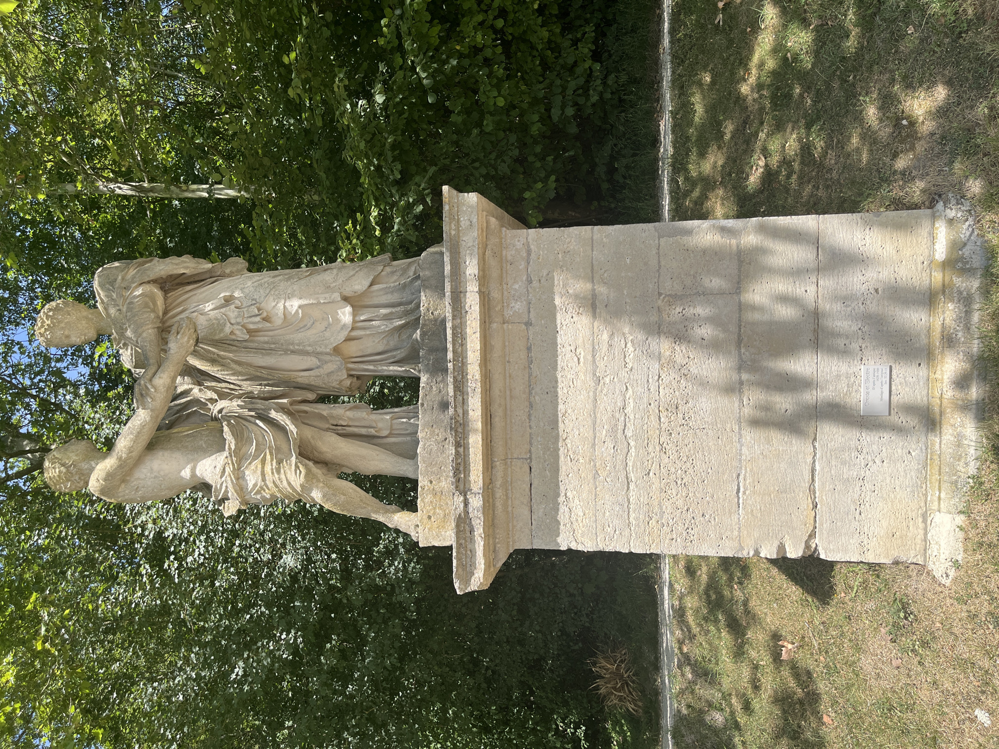 A stone statue of two figures, a male and a female, stands on a stone pedestal in a park. The male figure is to the left, depicted with a draped cloth around his waist and chest, reaching out his left arm towards the female figure. The female figure is to the right, wearing a draped garment and holding her arms towards the male figure. Both figures appear to be standing still. The pedestal is rectangular with a textured surface. Behind the statue, lush green trees form a dense backdrop. Sunlight casts shadows on the grass and the pedestal. A small plaque is affixed to the front of the pedestal. The statue is located in the Parc Frédéric Pic in Antony, France.