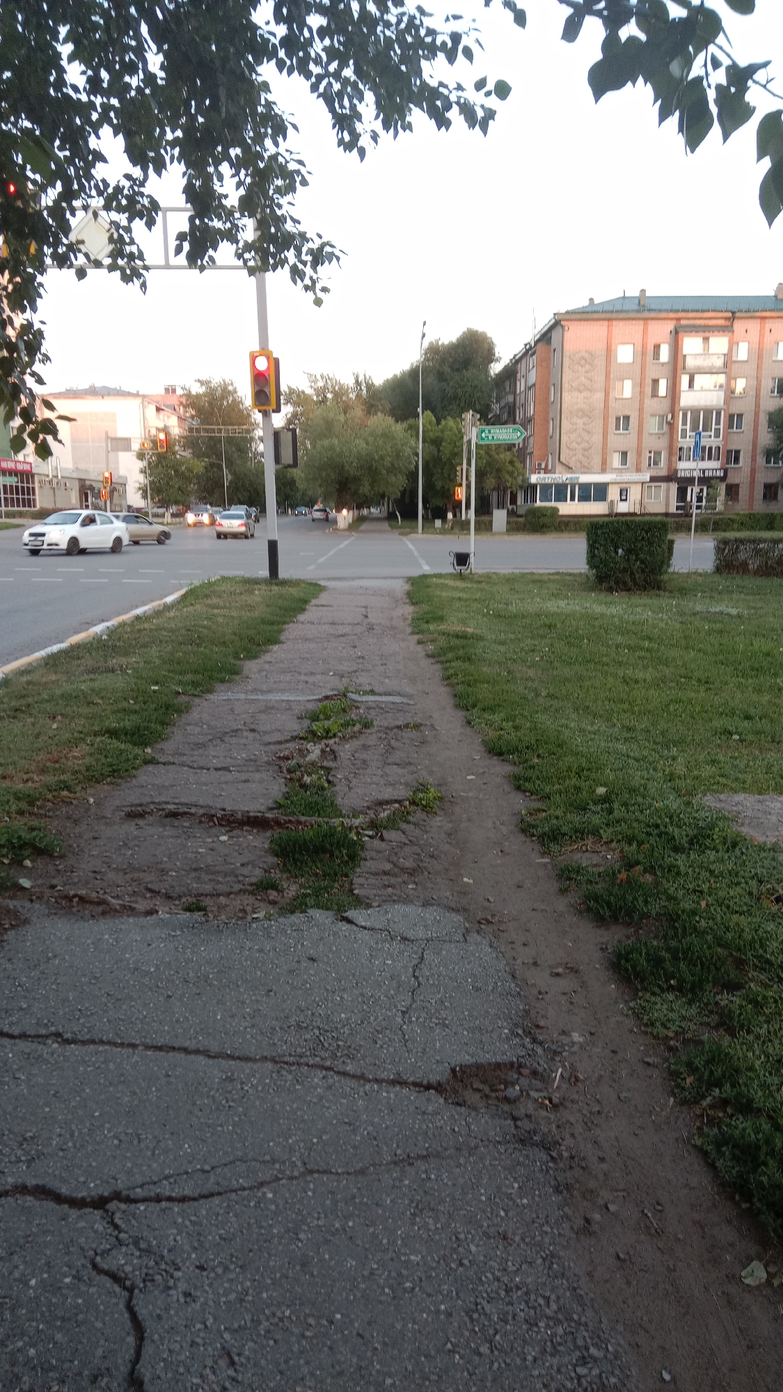 The image depicts a street scene in the city of Petropavl, Kazakhstan, likely during the daytime or twilight given the lighting conditions. A cracked and weathered concrete sidewalk occupies the foreground, bordered by a strip of grass. Beyond the sidewalk, a street is visible with several cars in motion. A traffic light, showing a red signal, is positioned on a pole to the left of the frame, indicating an intersection. In the background, apartment buildings line the street, with some displaying lights in their windows. Trees are scattered throughout the scene, with foliage partially obscuring the sky in the upper portion of the image. A street sign is visible on the right side of the intersection, providing directional information. The overall environment suggests a typical urban street setting.