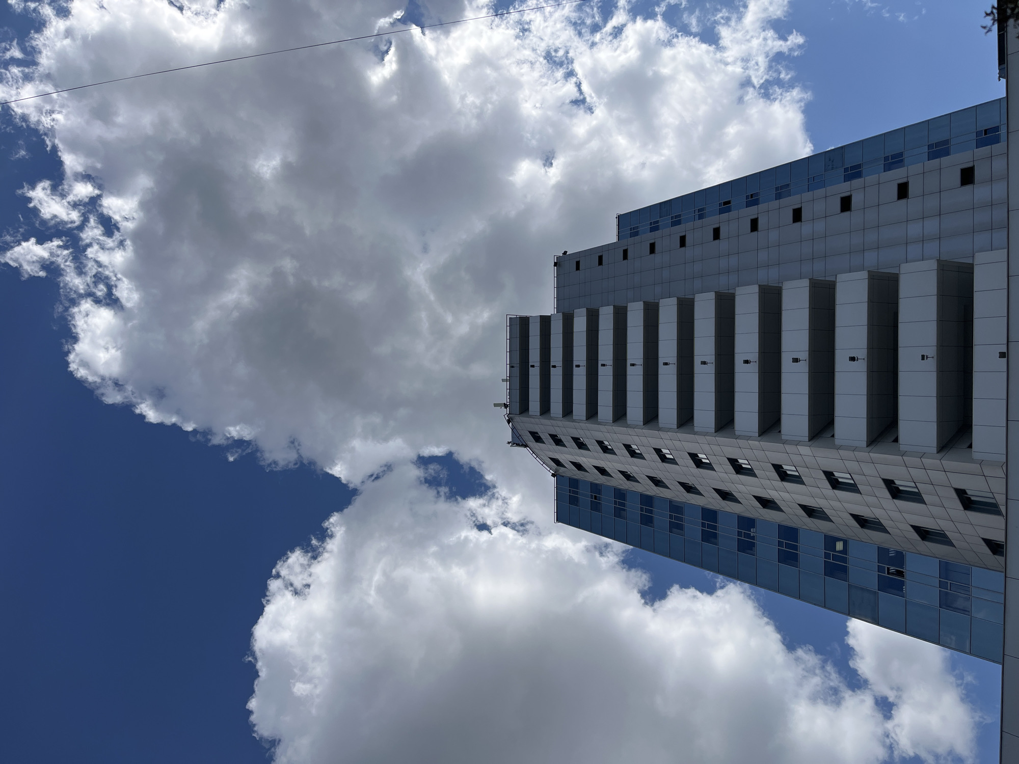 The image captures a low-angle view of a modern building against a backdrop of a partly cloudy sky. The building is characterized by a combination of solid, grey facade elements and reflective blue glass windows. The facade design incorporates projecting vertical rectangular structures. The sky alternates between patches of deep blue and fluffy white clouds. A thin wire is visible against the sky, suggesting an urban setting. While the location is stated to be Krasnoyarsk, Russia, no specific details in the image definitively confirm this.