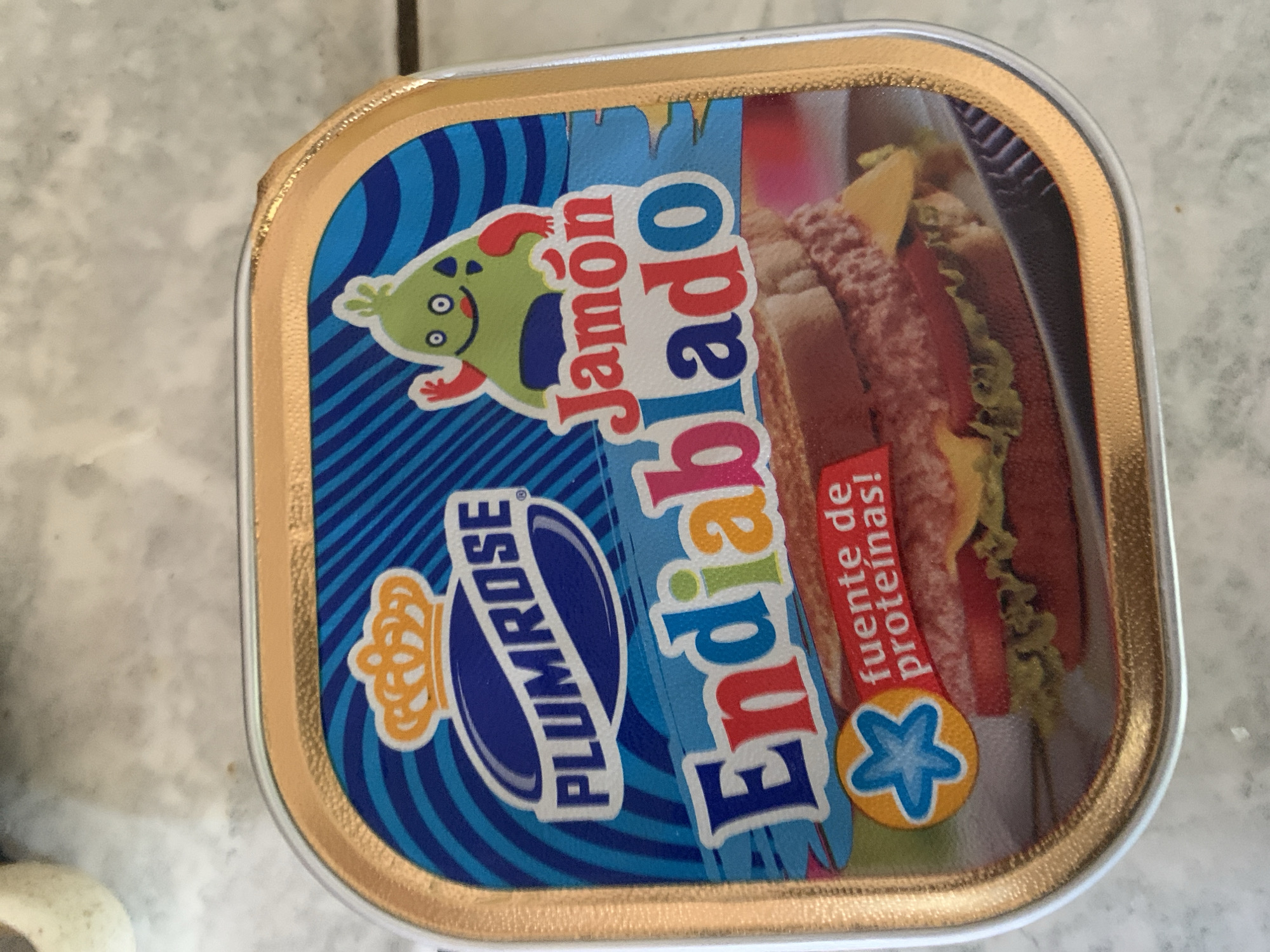 A rectangular, metallic food container, possibly a can or tray, is depicted in a high-angle shot, resting on a light-colored, textured surface that resembles a tiled countertop. The container's lid is adorned with a vibrant, predominantly blue label featuring diagonal stripes in various shades of blue.
The label showcases a cartoonish green character with red limbs and a smiling face, positioned above the word "Jamón" rendered in stylized red lettering. Below this, the word "Endiablado" is prominently displayed in large, multi-colored letters, suggesting the product is "Deviled Ham." To the left, a distinct blue oval emblem with a golden crown prominently features the brand name "PLUMROSE" in white capital letters.
On the right side of the label, a clear image of a sandwich or hotdog is visible, containing what appears to be processed meat, fresh lettuce, and cheese, illustrating the product's potential use. A red ribbon-like banner partially covers this food image, bearing the white text "fuente de proteínas!" (source of protein!). A small, circular orange emblem with a blue star design is also visible near the bottom right corner of the label.
The container itself has a noticeable golden or bronze-colored rim with a corrugated texture around its edges. The lighting suggests an indoor setting during the daytime. No individuals are present, and the image solely focuses on the packaged food item.