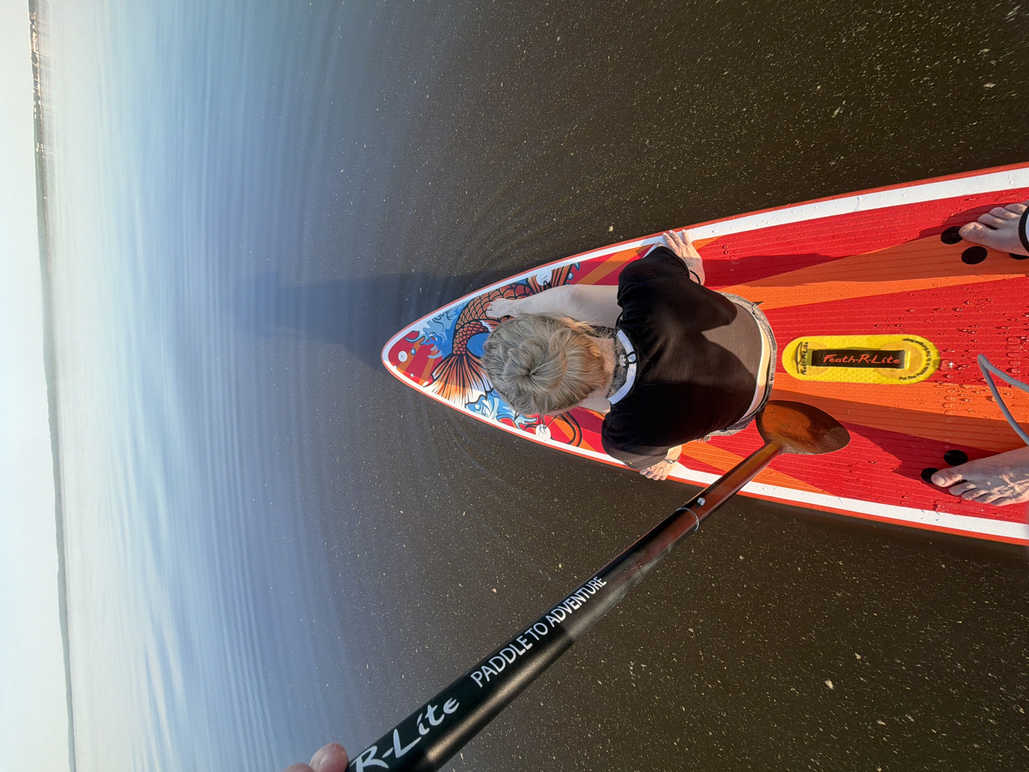 A high-angle shot shows a person on a paddleboard in a body of water. The paddleboard is predominantly orange with red and white borders, and a decorative image of a fish is visible at the board's nose. The person is kneeling on the board, wearing a black shirt. A wooden paddle rests horizontally across the board. A black paddle with white lettering reading "R-Lite PADDLE TO ADVENTURE" is also present. Feet are visible near the back of the board. The surrounding water appears calm, and a shoreline is visible in the distance. There is no discernible information from the image to associate the image with the city of Noviy, Russia.