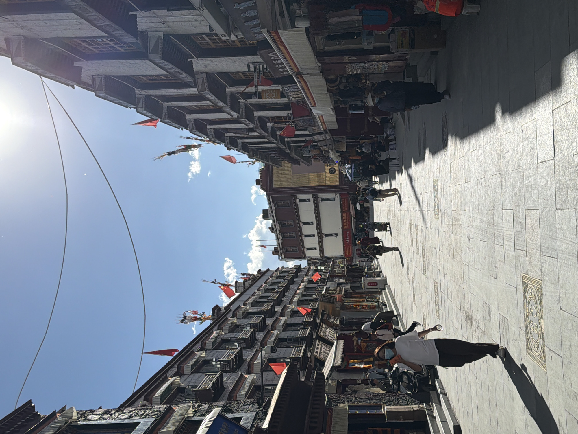 The image displays a lively street scene in Chengguan District, China, under a bright, sunny sky with a few scattered clouds. The street is wide and paved with light-colored tiles, featuring decorative patterns and what appears to be a stylized golden symbol or script, possibly Tibetan, embedded into the ground.
The street is lined on both sides by multi-story buildings exhibiting traditional architecture, characterized by dark wooden elements, intricate window frames, and projecting balconies. Numerous red flags, some resembling the national flag of China, are mounted on the building facades, and colorful prayer flags are also visible, particularly towards the left side of the frame. Shop awnings extend over some storefronts, and goods appear to be displayed outside.
Several people are present on the street. In the foreground, a person wearing a white t-shirt, dark pants, sneakers, and a blue face mask is walking, holding a small plastic bottle. Long, distinct shadows are cast on the pavement, indicating strong, direct sunlight from a high angle, suggesting it is mid-morning or afternoon. Further down the street, other individuals are seen walking or standing near the shops, engaging in typical street activities such as shopping or sightseeing. Overhead, thin cables or power lines traverse the blue sky.
The scene depicts a bustling cultural or commercial district. No specific text on shop signs is clearly legible.
