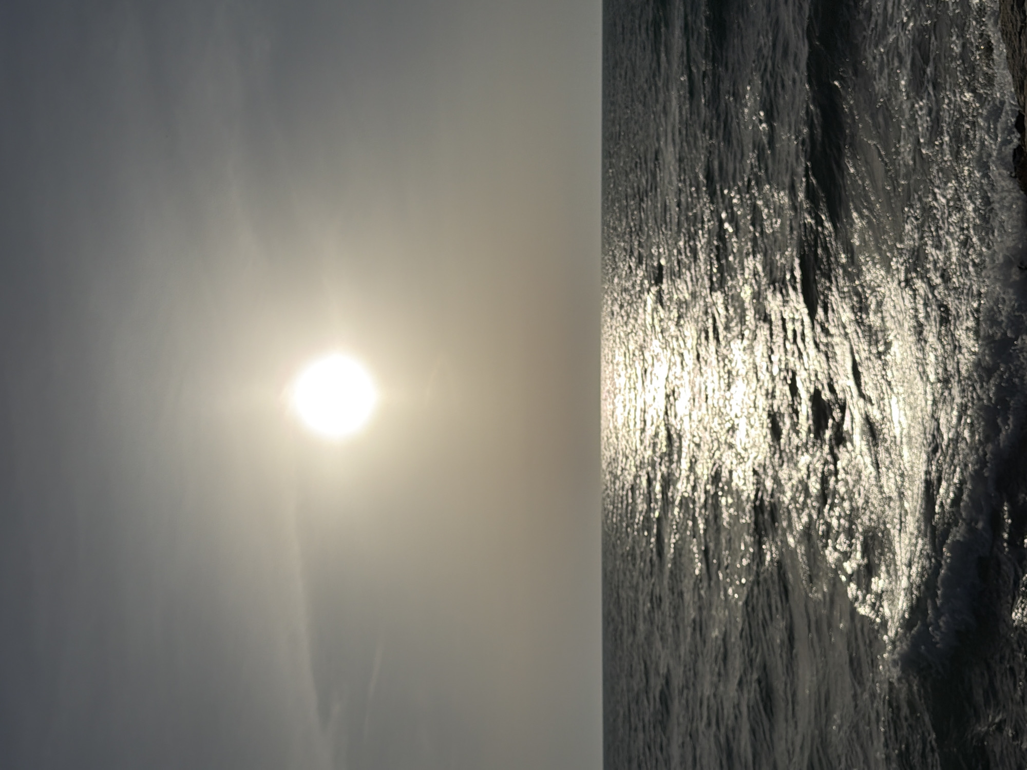 The image shows a seascape in Tel-Aviv, Israel. The sea, reflecting the sun, is on the right, and a hazy sky with the sun is on the left.