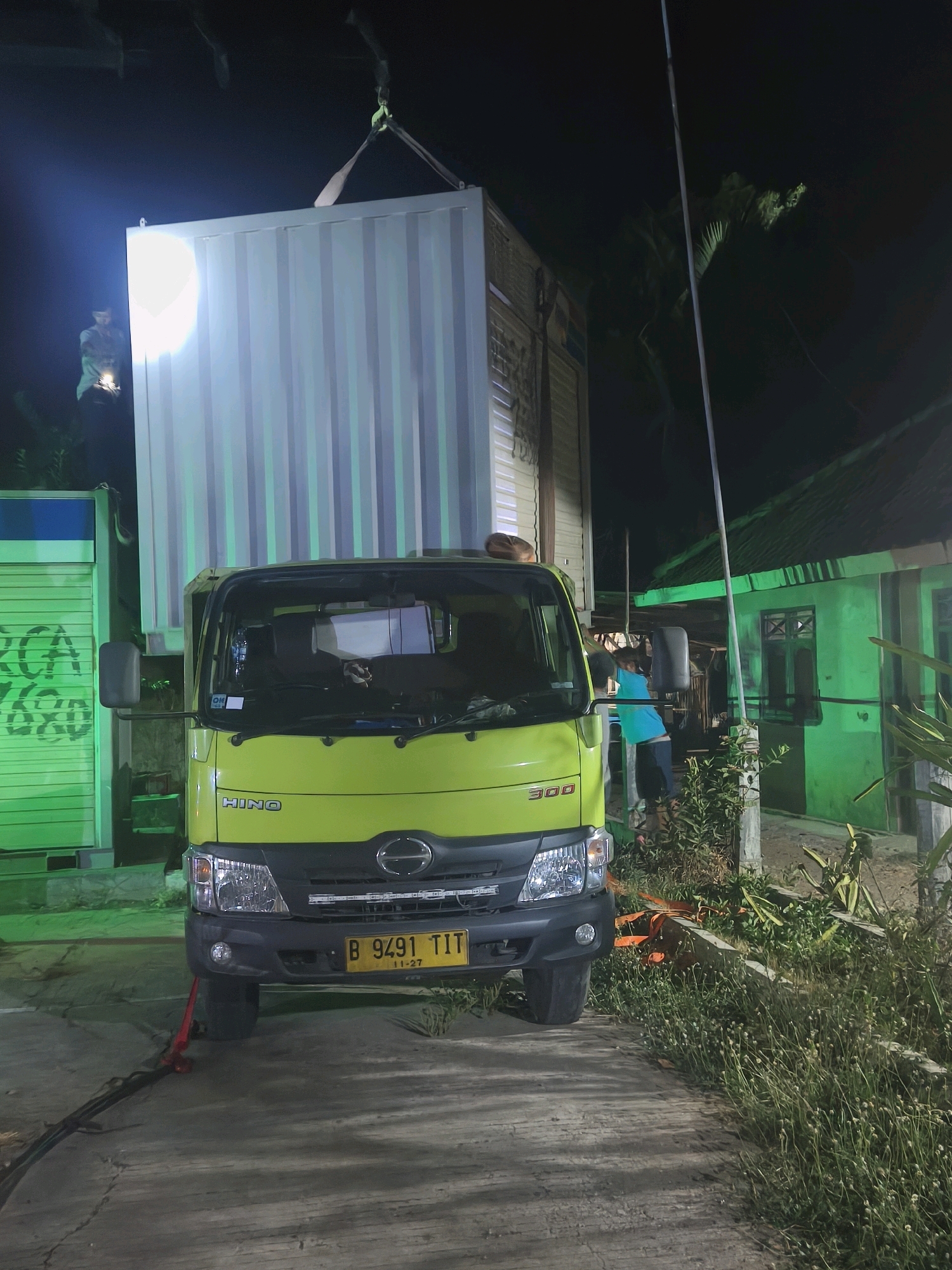 In the city of Kroya, Indonesia, at night, a bright green Hino truck is parked on a concrete surface, partially supported by a red hydraulic jack. The truck's license plate reads "B 9491 TIT 11-27". A large, off-white shipping container is suspended above the truck by a crane. In the background, a green building with a corrugated roof and a window is visible. To the left of the truck, another green structure with a roll-up door and blue and white markings is present. A person in a turquoise shirt and dark shorts is standing near the side of the truck, facing away from the camera. Another person, holding a bright light source, stands to the left of the suspended container, partially illuminated by its glow. The scene is illuminated by artificial lights, creating strong contrasts and shadows. Vegetation, including grass and plants, lines the right side of the image.