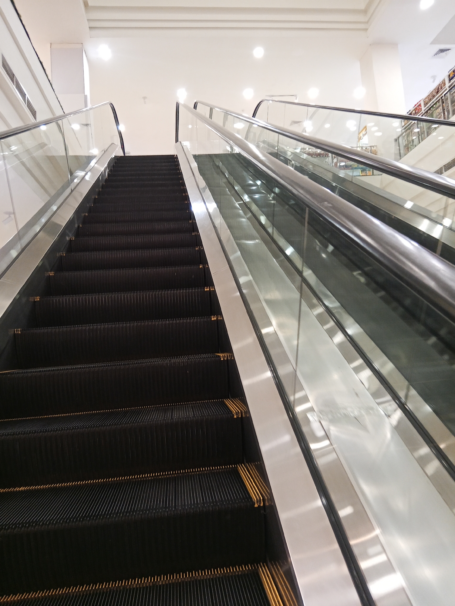 An upward-angled view shows a commercial escalator with black treads and metallic gold trim, flanked by glass and stainless steel railings. The escalator ascends into a brightly lit interior space with a white ceiling and recessed lighting. The context provided suggests this scene is located in Lubuklinggau, Indonesia, likely within a shopping mall or similar commercial establishment. There are no people visible in the frame.