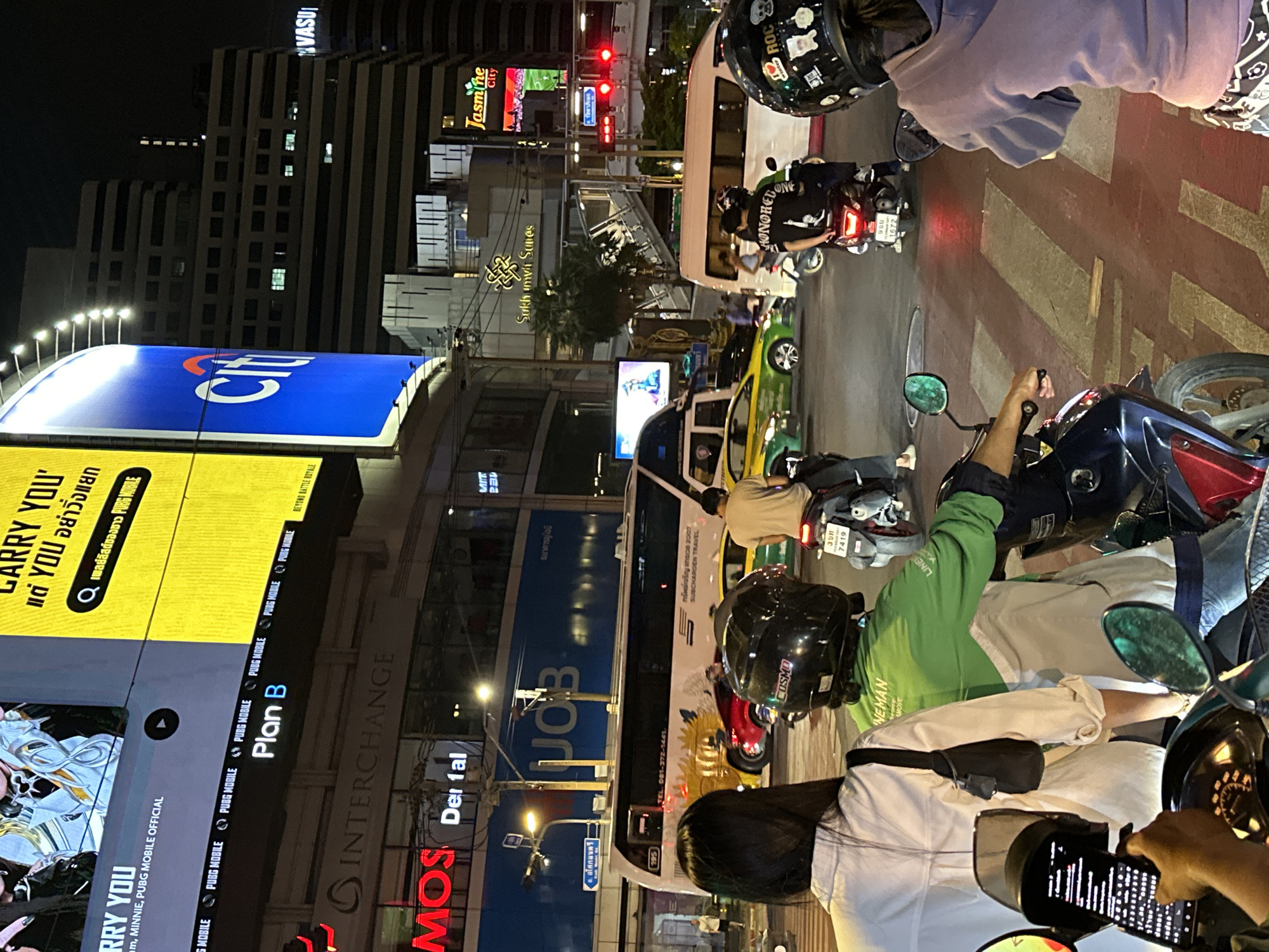 The image depicts a nighttime street scene in Bangkok, Thailand, viewed from the perspective of someone on a motorcycle. Multiple motorcycles are positioned at a red light, indicating a pause in traffic. A large electronic billboard displays advertisements, including one for PUBG Mobile, and signage for various businesses such as "Interchange," "MOS Dental," "UOB Bank" ("ธนาคารยูโอบี"), and "Sukhumvit Suites." A white passenger van is among the stopped vehicles. Pedestrians and other traffic are visible in the background, along with buildings and street lights. The Jasmine City building is visible in the distance. Helmets on motorcyclists have stickers on them. The photo is rotated 90 degrees to the left.