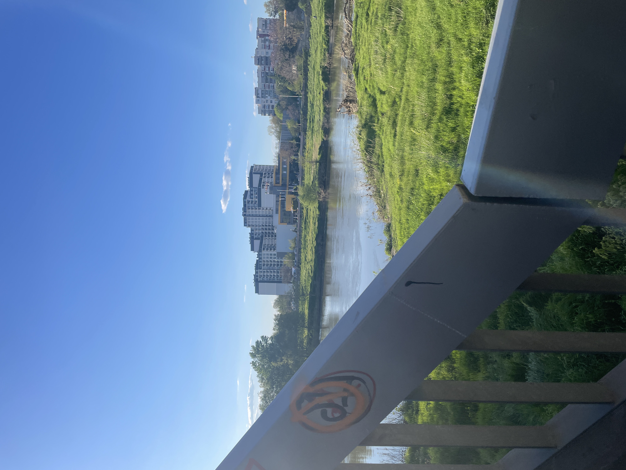 The image captures a sunlit urban landscape in Plovdiv, Bulgaria, featuring a river flowing through a developed area. The scene is viewed from an elevated position, likely a bridge or walkway, with a substantial gray metal railing in the foreground, partially obscuring the lower right portion of the view. On this railing, an orange and black graffiti tag, resembling a stylized "16" or an abstract symbol, is visible. A subtle lens flare or rainbow effect can be seen on the railing to the far right.
The river runs through the midground, its surface reflecting the clear blue sky above. Both banks of the river are covered in lush, vibrant green grass. Along the left bank, a series of modern, multi-story apartment buildings with varying architectural styles line the waterfront, interspersed with trees. The right bank is also covered in green grass and some scattered trees in the distance.
The sky is a bright, clear blue with a few small, wispy white clouds visible towards the right side. The strong illumination suggests it is daytime, likely morning or early afternoon, under sunny weather conditions. There are no human figures or specific activities depicted, conveying a sense of calm and a harmonious blend of urban development and natural elements.