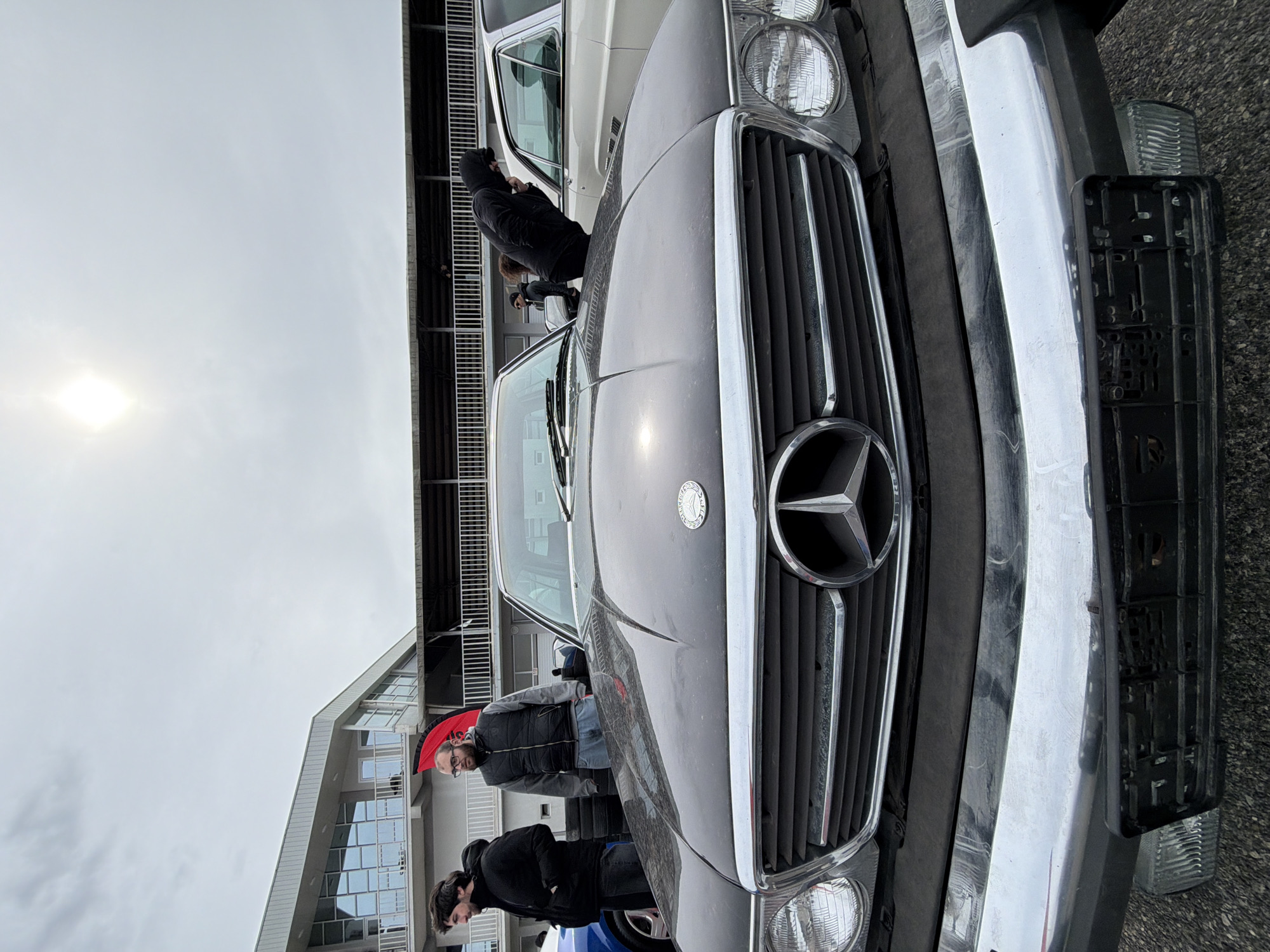 A low-angle, full-color shot features the front end of a dark-colored Mercedes-Benz, likely a vintage model, with its distinctive chrome grill and star emblem prominently displayed. To the right of the car, a chrome bumper and partially visible license plate frame are noticeable.
Two individuals are partially visible in the background, positioned to the left of the vehicle's hood. One is wearing a dark jacket and jeans. Behind them, a building with a modern architectural style featuring glass windows and a light-colored facade is visible. A structure resembling bleachers or a tiered platform is present behind the building. The sky is overcast, with the sun visible as a bright spot.
The location is potentially in Algetis Meurneoba, Georgia.