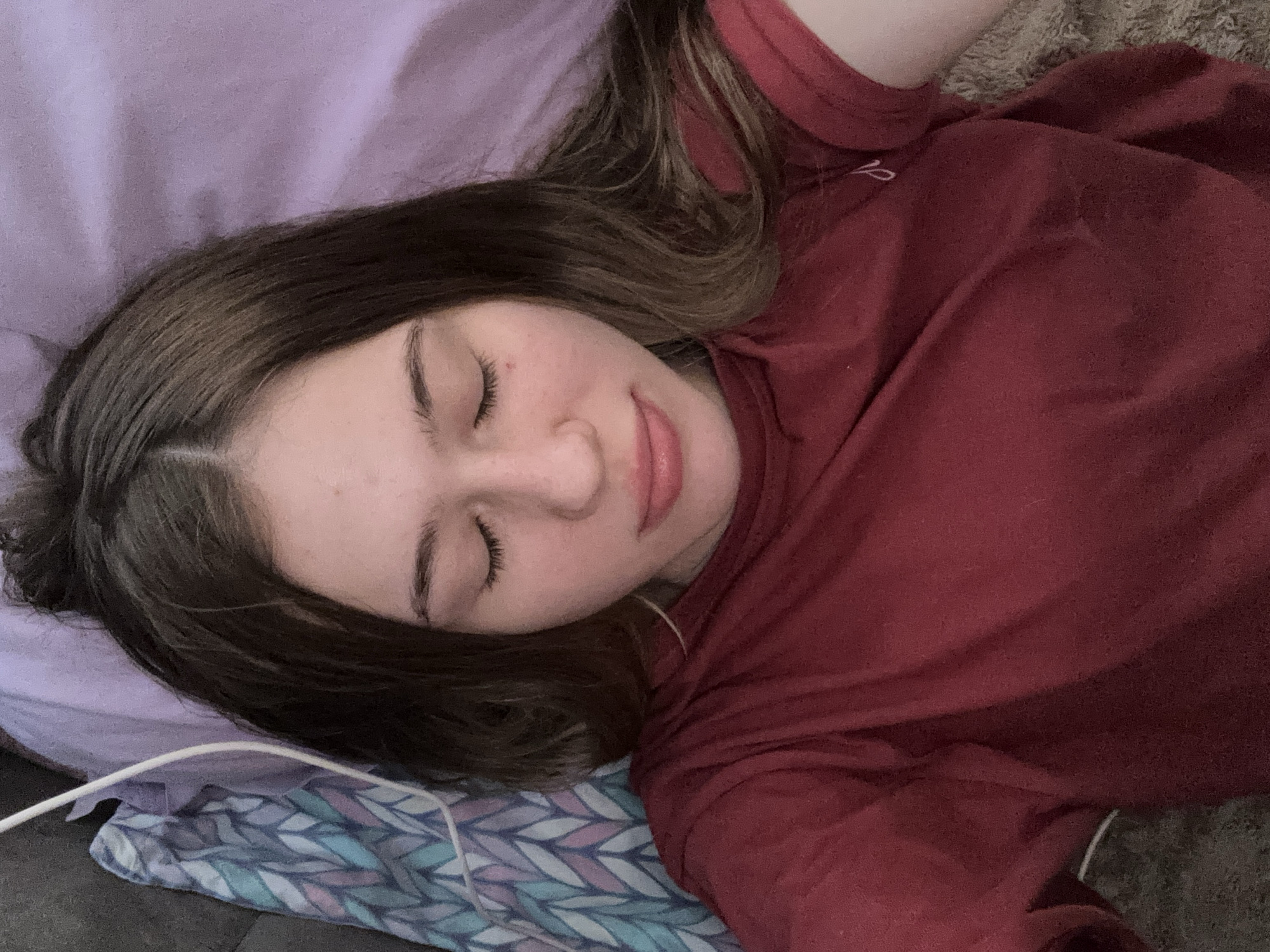 A young woman with brown hair and closed eyes lies on a bed, her head tilted slightly to the right. She is wearing a dark red t-shirt. To her left, a purple pillow and a patterned blanket are visible. The environment appears to be a bedroom. The image was taken in the city of Sinyavino, Russia.